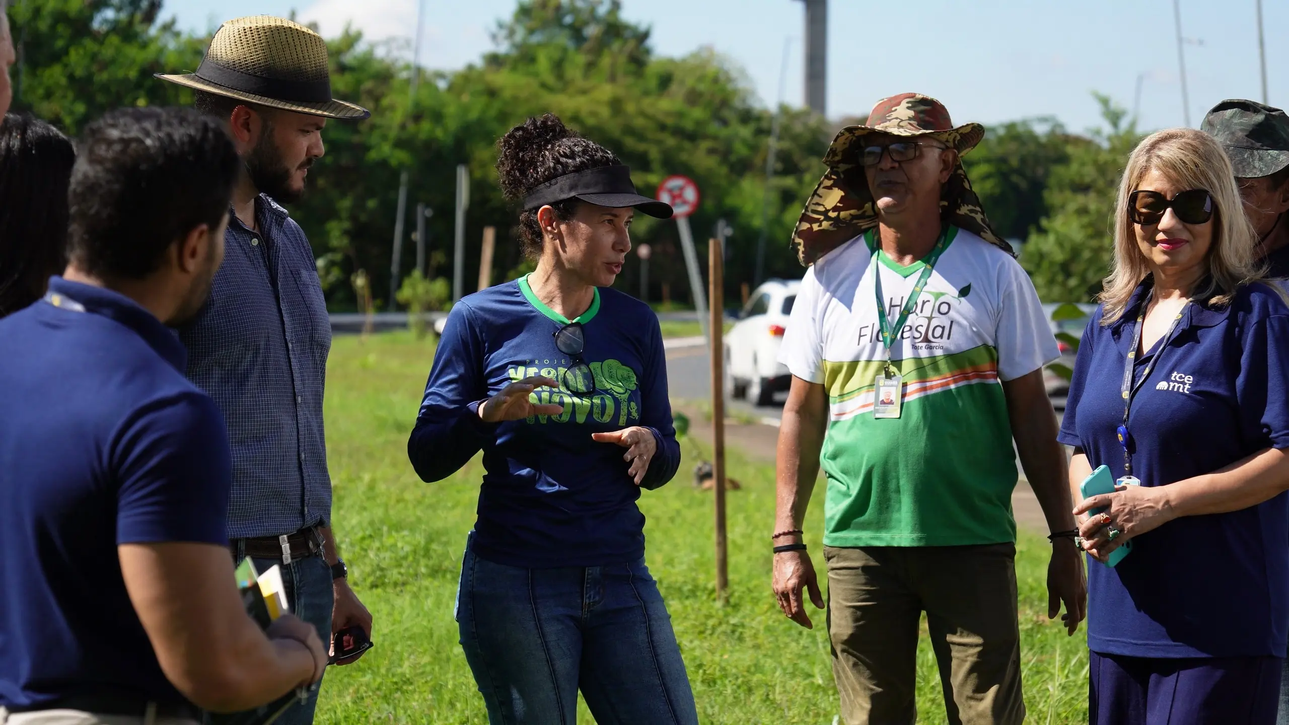 Um grupo de cinco pessoas em pé em um gramado conversando. Uma mulher de boné e blusa azul com logo "Mato Grosso" fala para as outras. Ao seu lado, um homem de chapéu camuflado e camisa verde/branca com logo "Horta Florestal". Outras pessoas observam.