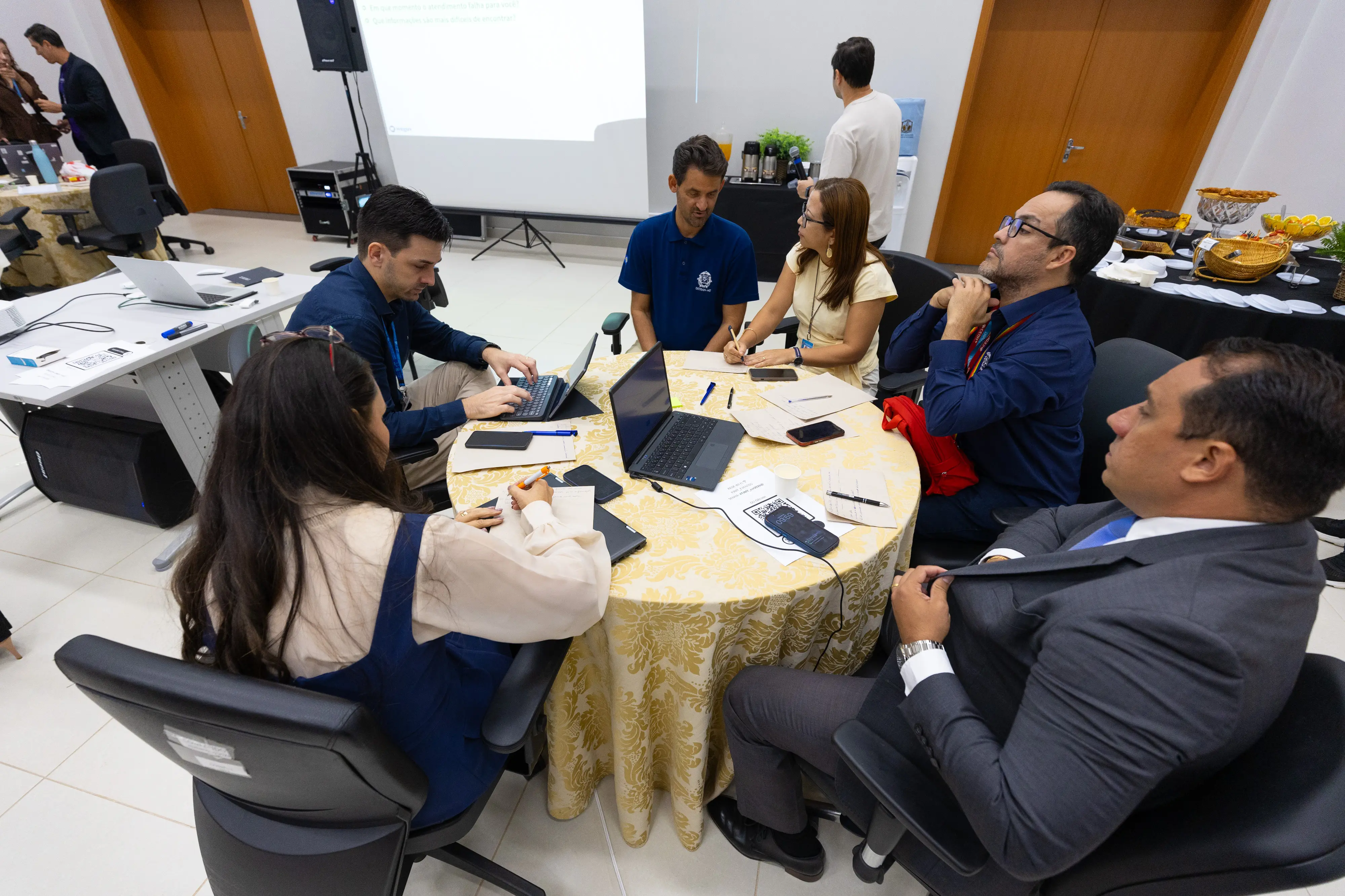 Participantes discutem propostas em roda de conversa durante oficina de planejamento. Ambiente possui mesas circulares.