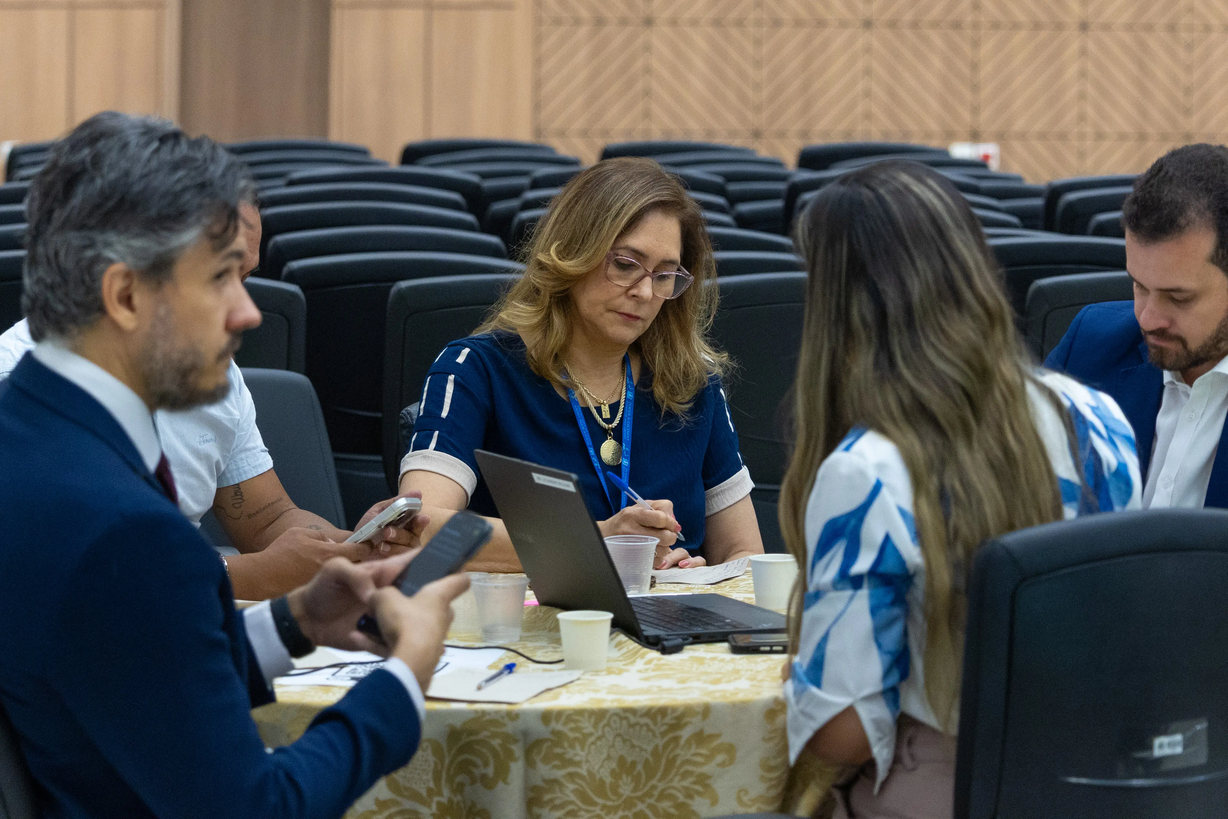 Grupo reunido em mesa redonda participa de atividade colaborativa. Pessoas conversam e anotam ideias durante oficina.