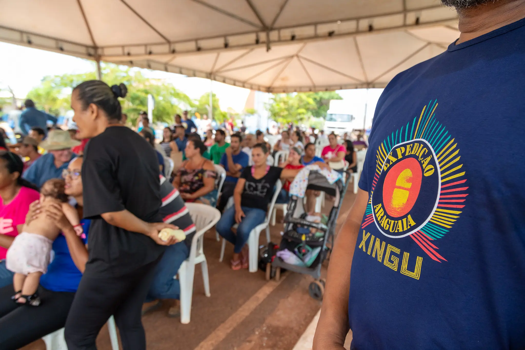 Em primeiro plano, no canto direito, parte da camisa na cor azul do projeto Expedição Araguaia-Xingu. Ao fundo, desfocado, pessoas sentadas em cadeiras, aguardando na fila de atendimento.