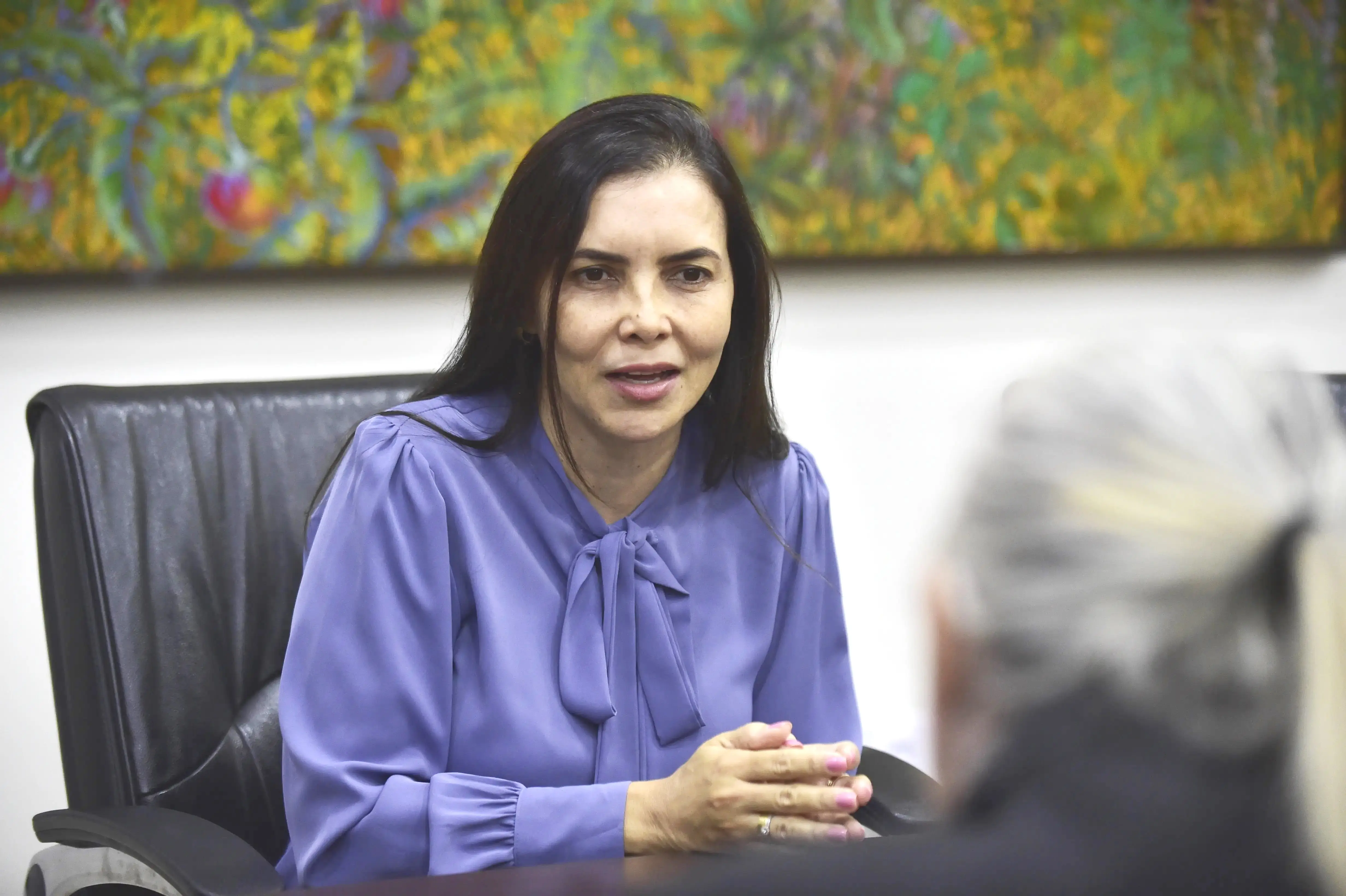 Juíza Anna Paula é uma mulher da pele clara, cabelos lisos. Ela veste uma blusa azul claro. Ela está sentada em uma cadeira com estofado preto e suas mãos estão sobre uma mesa.