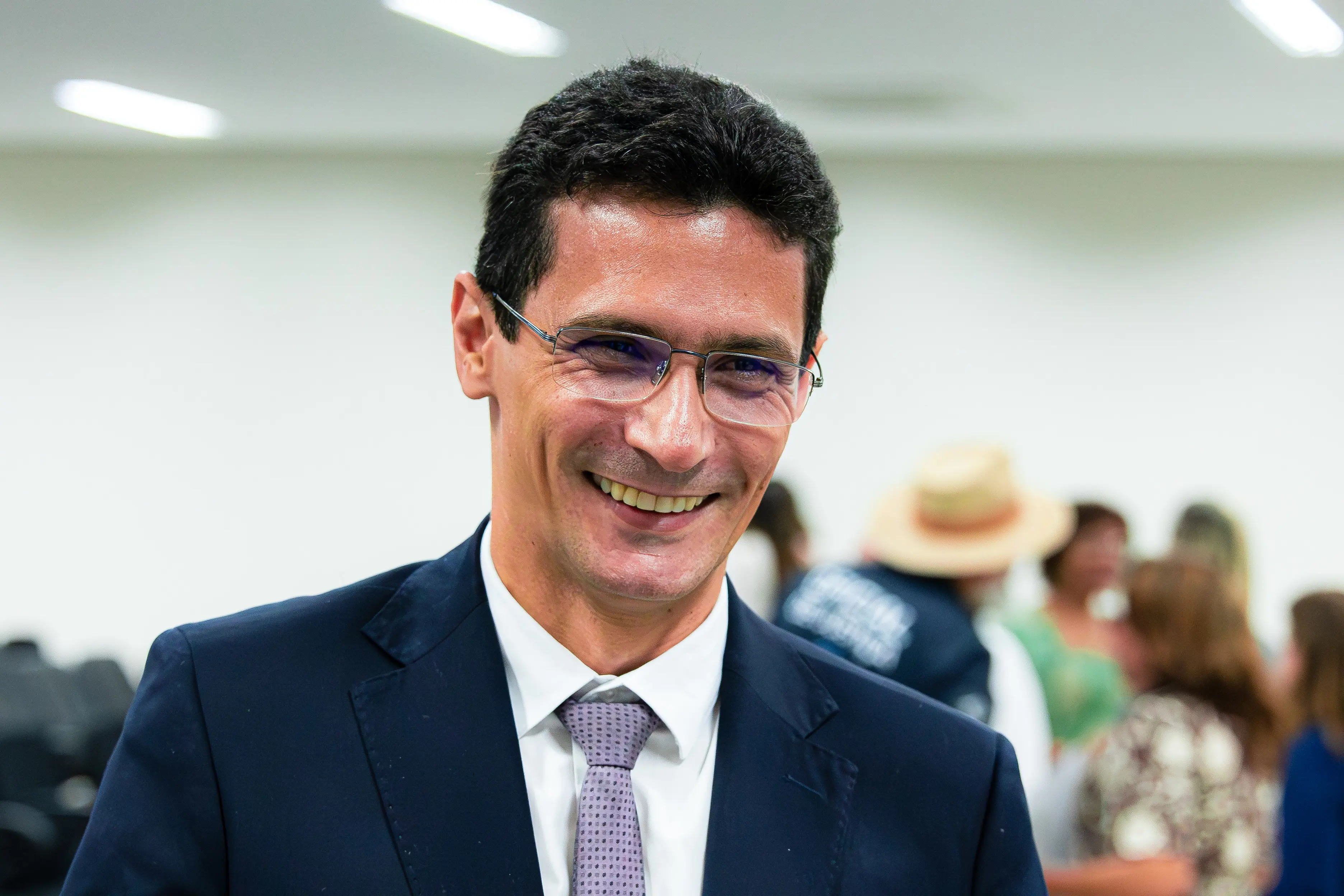 Homem sorri amplamente vestindo terno escuro, camisa branca e gravata. Usa óculos de grau e está em um ambiente interno com pessoas desfocadas ao fundo.