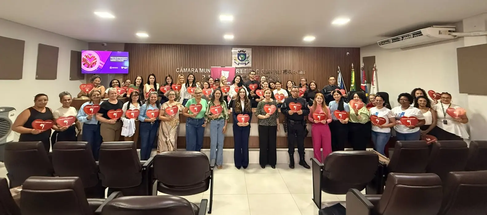 Foto horizontal que mostra cerca de 50 pessoas em pé em um auditório. elas posam para a foto segurando panfletos vermelhos em formato de coração. 