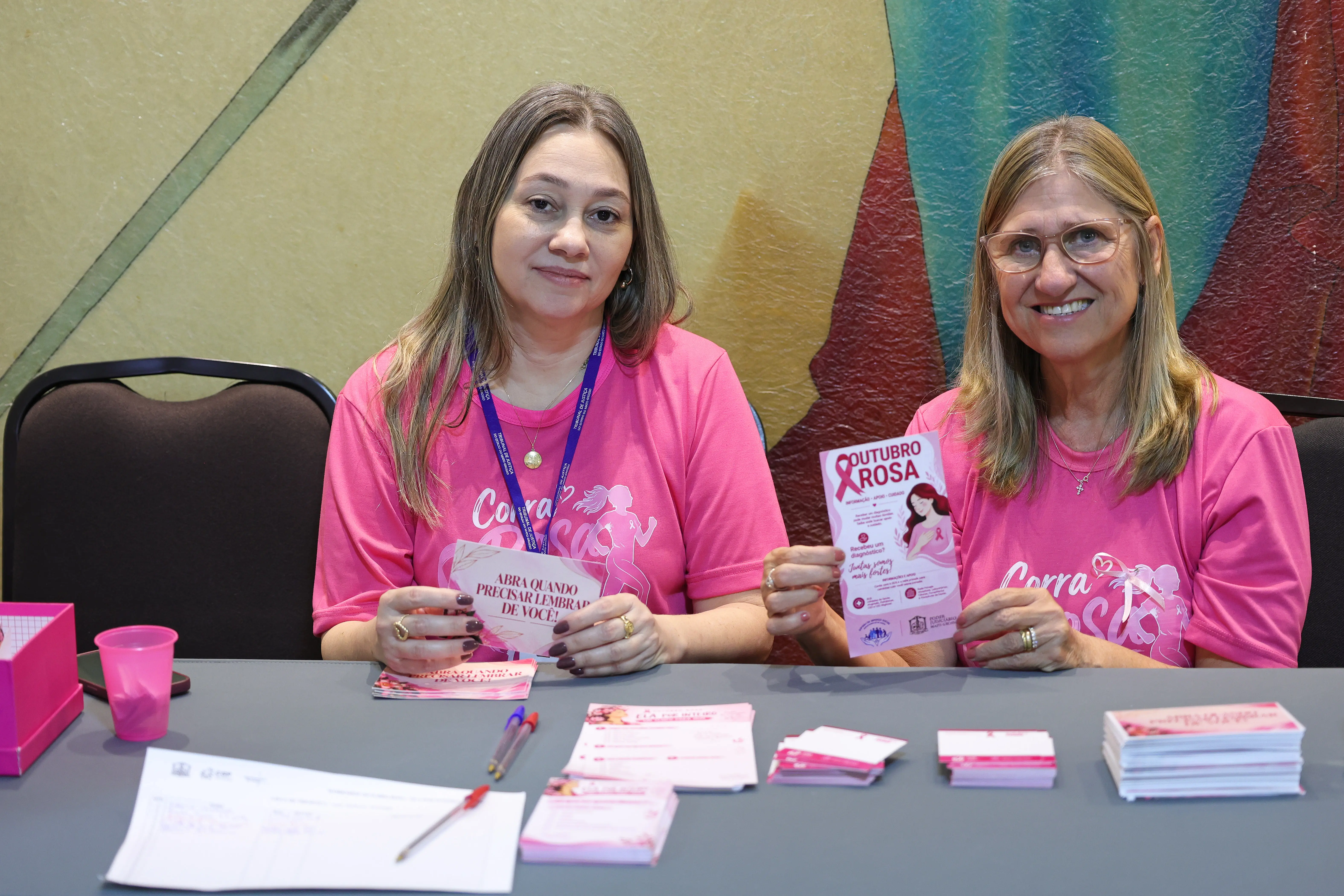 Duas mulheres de rosa atendem participantes à mesa no espaço “Um tempo para mim”; duas delas fazem anotações. Em primeiro plano, cartazes da ação.