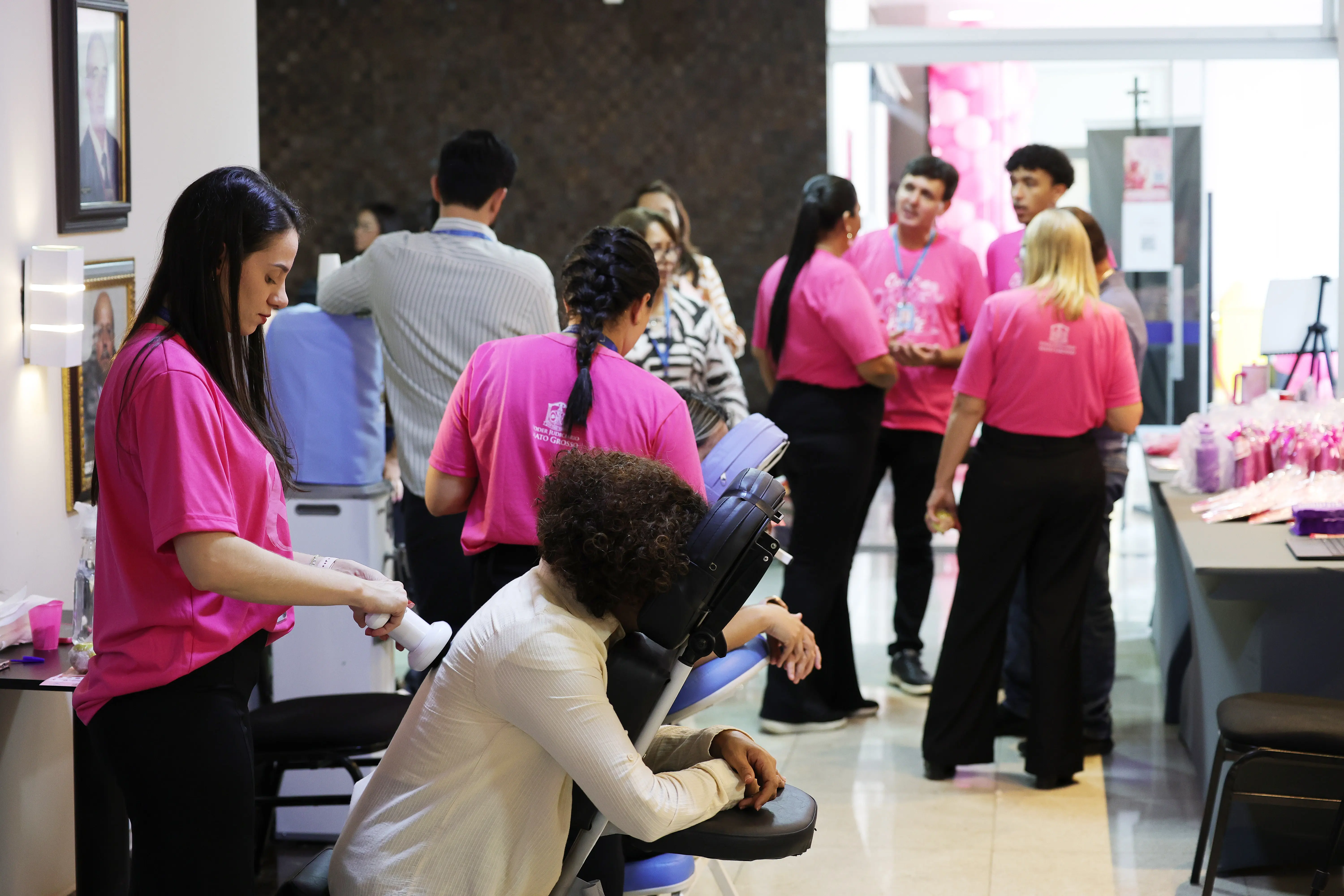 Mulher de camiseta rosa faz massagem com aparelho nas costas de servidora sentada em cadeira de massagem; ao fundo, participantes conversam no hall do Tribunal.