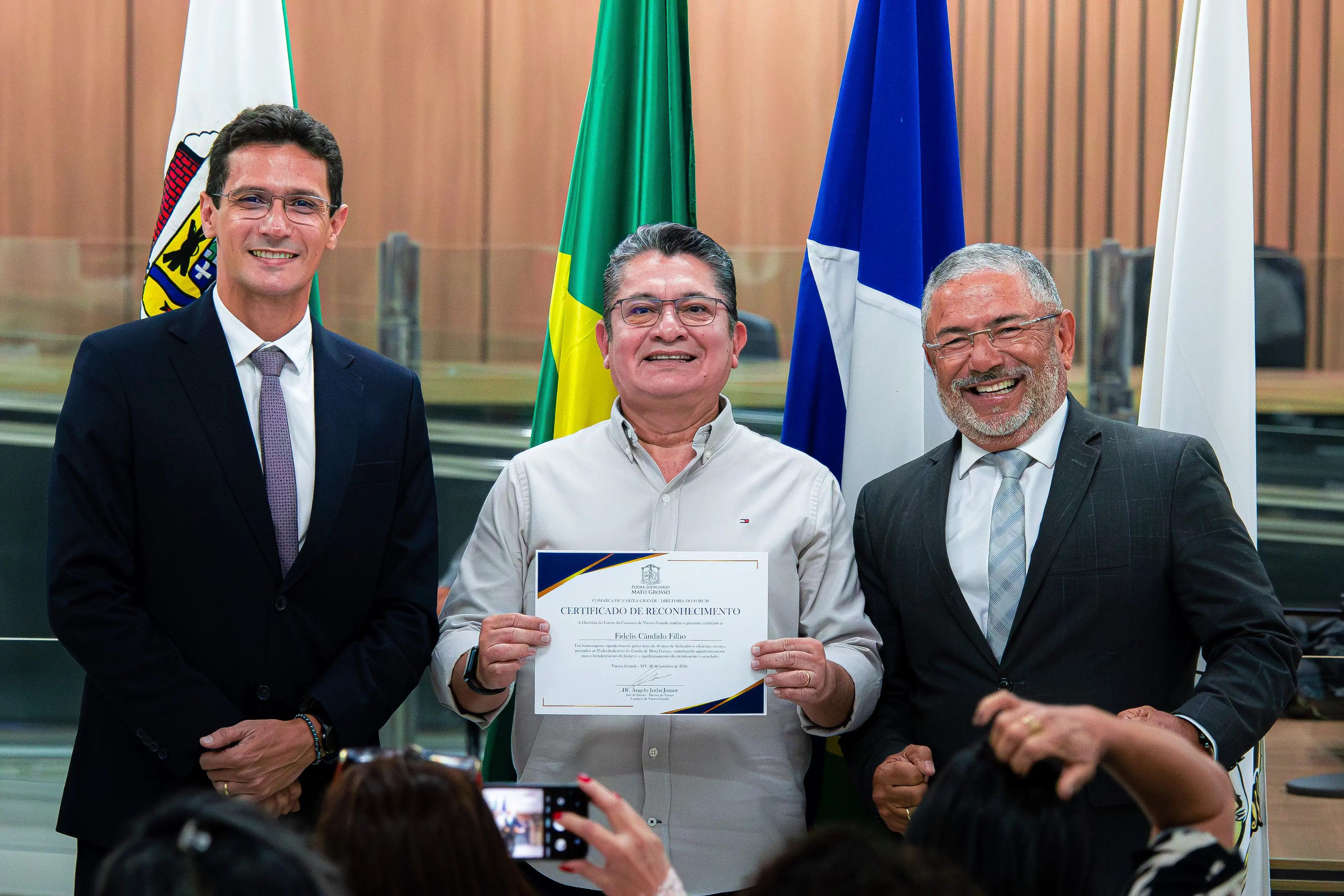 Três homens sorriem ao centro. No meio, um homem de óculos segura um certificado de reconhecimento. Ao lado dele, dois homens de terno e gravata. Ao fundo, bandeiras institucionais.