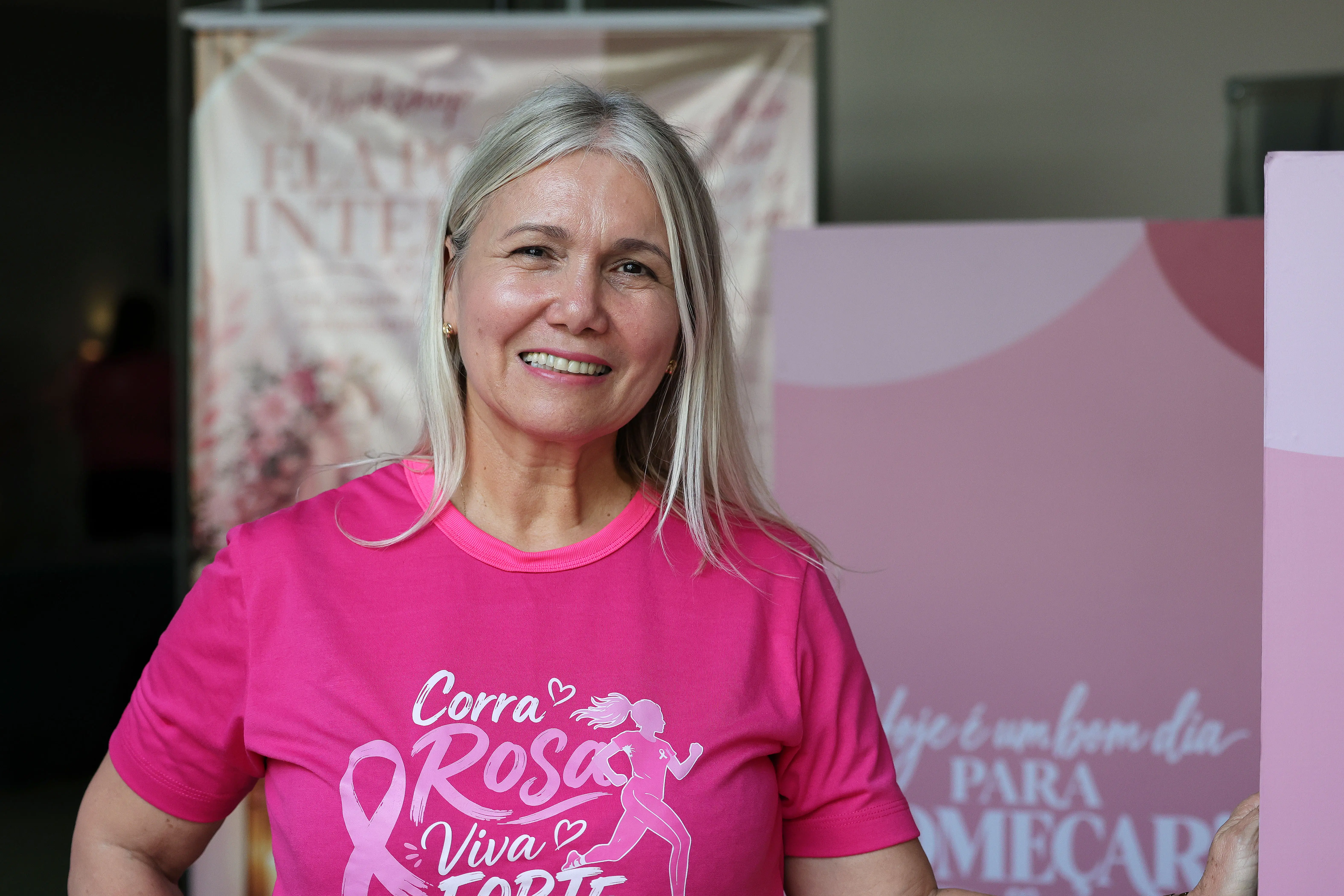 Mulher de cabelos loiros e camiseta rosa sorri em frente a banner do workshop Ela por Inteiro e painel rosa, durante o evento do Outubro Rosa.