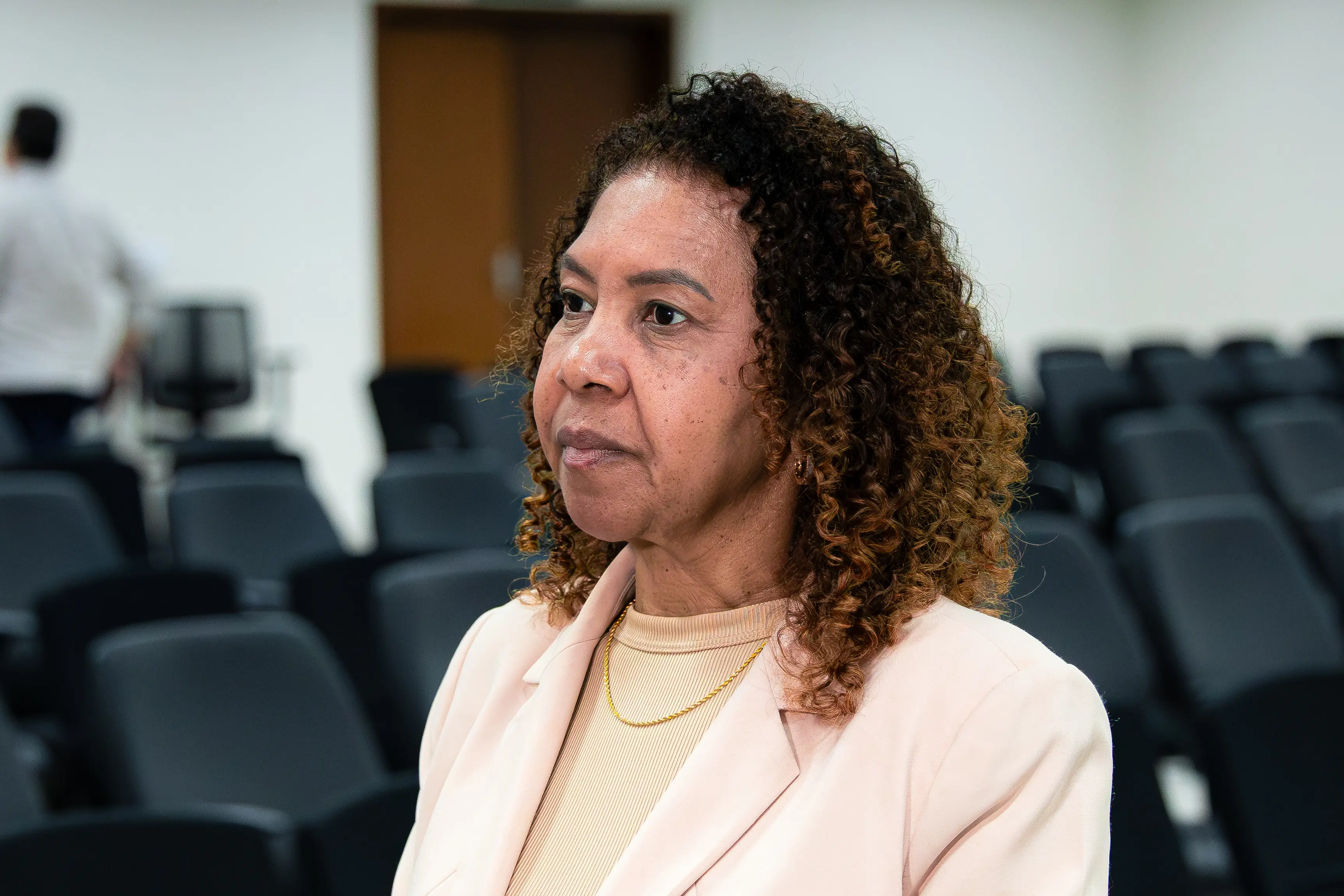 Mulher negra de cabelos cacheados médios olha atenta para o lado. Veste blazer em tom claro e blusa de gola alta. Ao fundo, auditório com fileiras de cadeiras escuras.