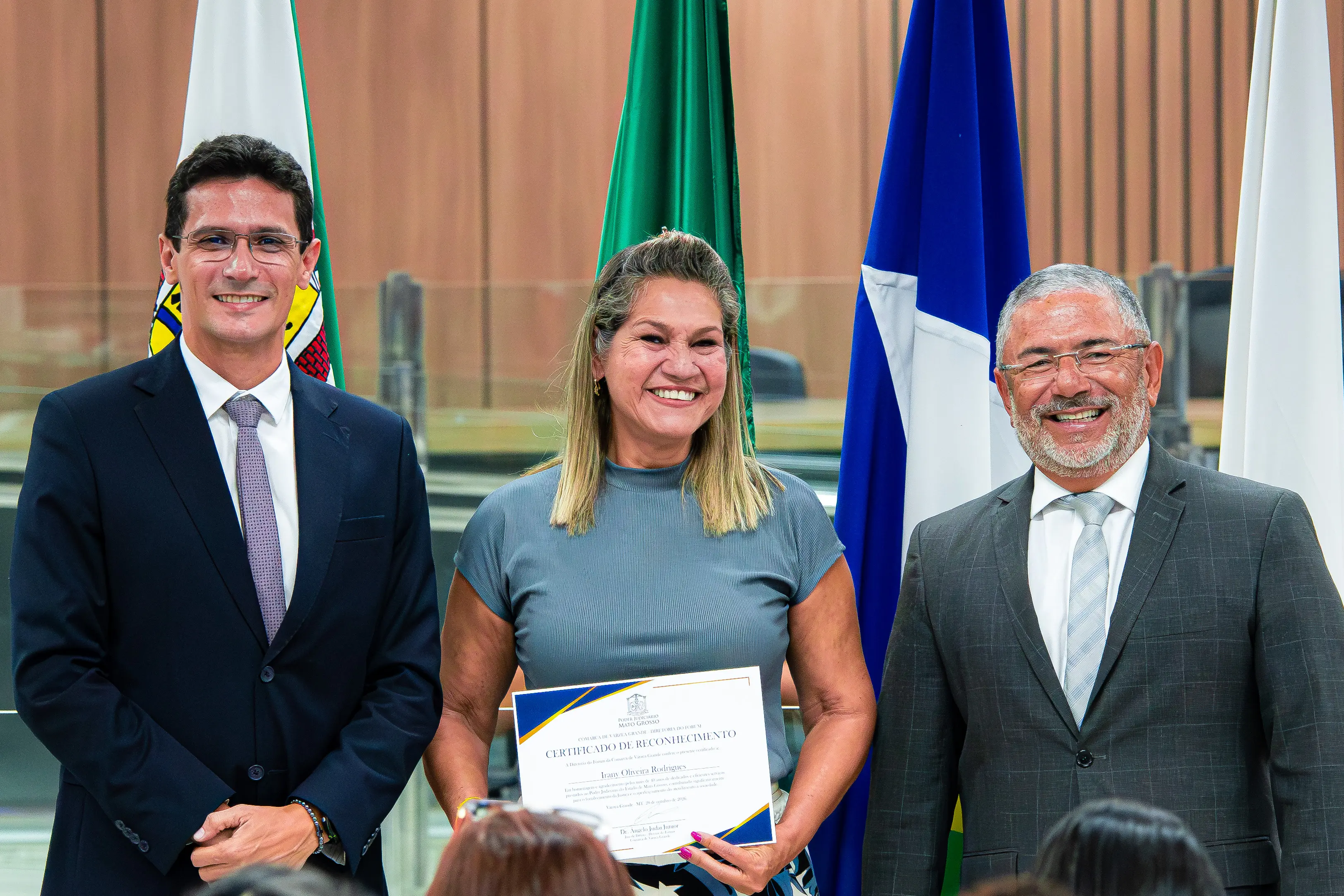 Três pessoas sorriem ao centro. No meio, uma mulher segura um certificado de reconhecimento. Ao lado dela, dois homens de terno e gravata. Ao fundo, bandeiras institucionais.