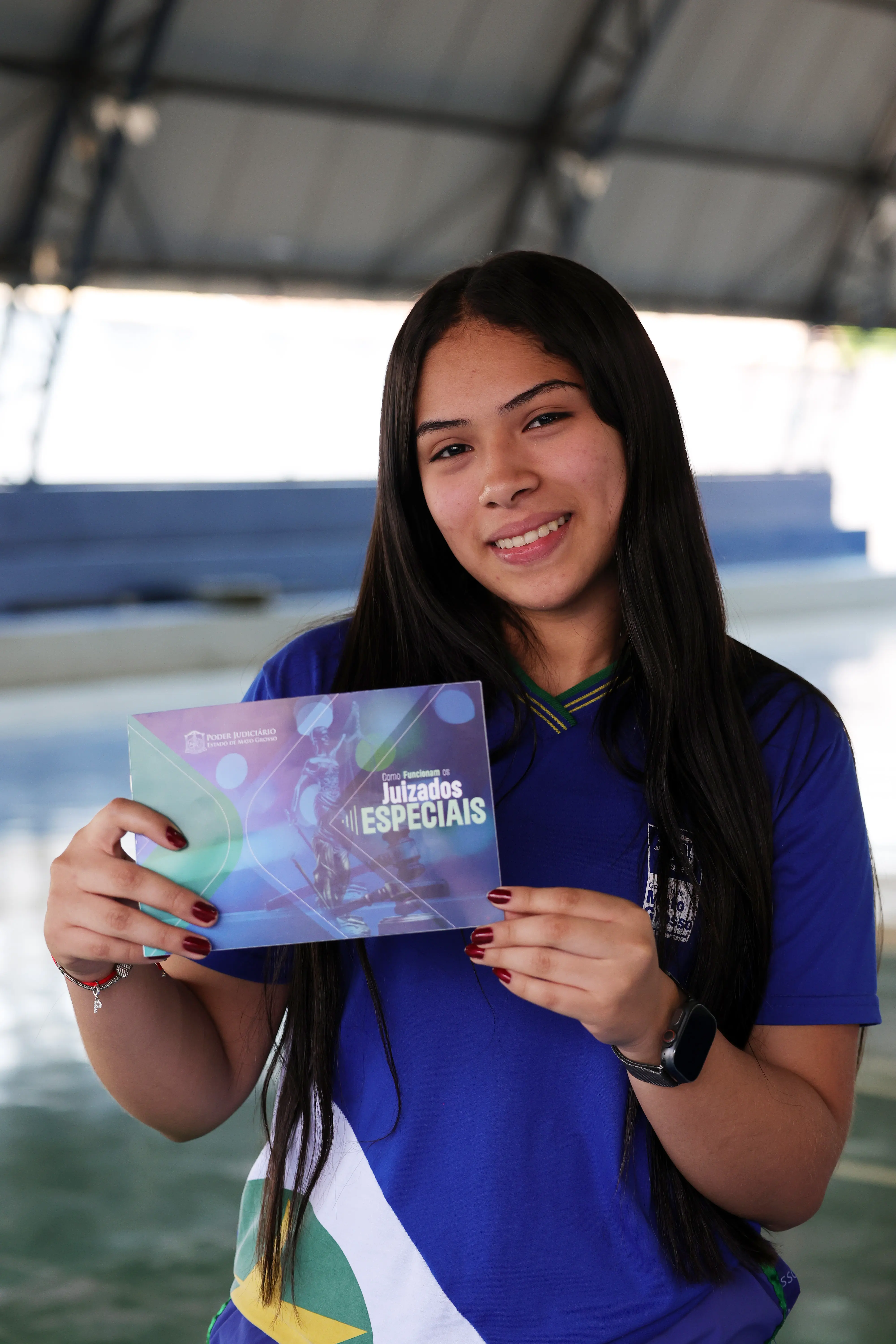 Uma adolescente com cabelos longos e escuros sorri para a câmera, vestindo uma blusa azul de uniforme escolar, enquanto segura com as duas mãos um livreto com a inscrição "Como Funcionam os Juizados Especiais" na capa.