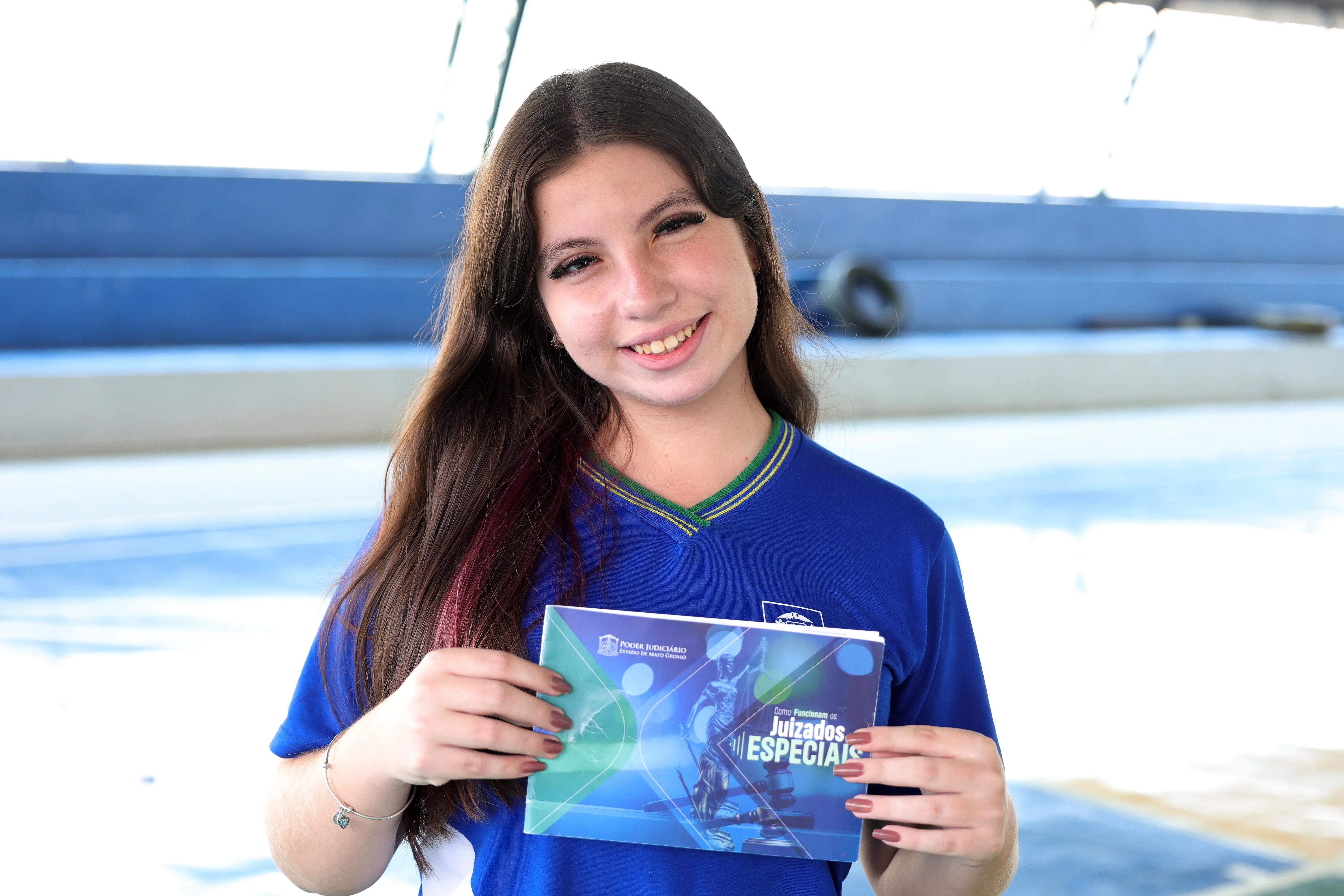 Uma jovem de cabelos longos e castanhos sorri levemente, vestindo uma camisa azul de uniforme com gola verde e amarela, e segura com as mãos um livreto aberto com o título "Como Funcionam os Juizados Especiais".