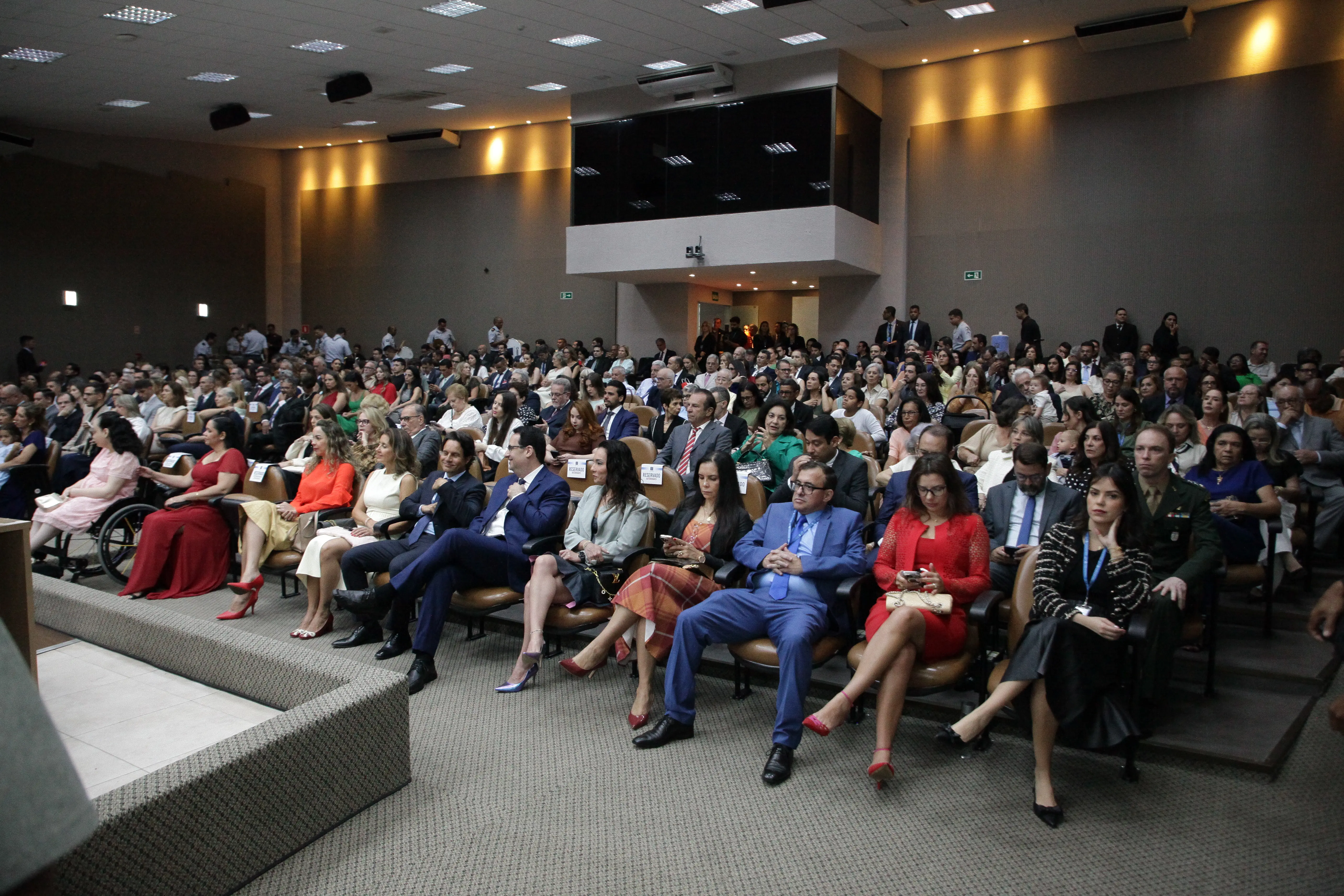 Auditório completo em evento solene. Público elegante ocupa todas as poltronas. 