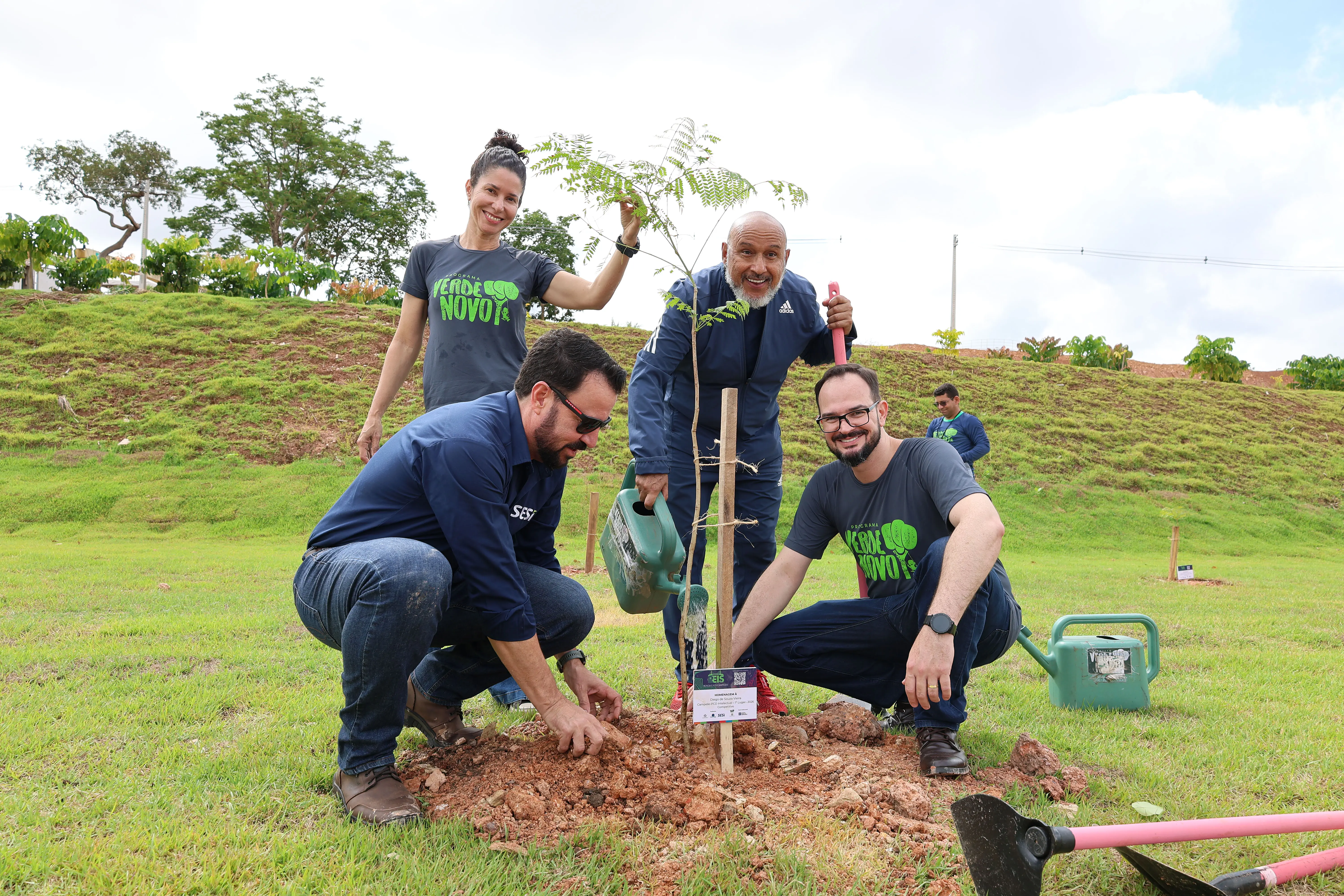 Quatro pessoas sorridentes plantam uma muda de árvore em um gramado. Dois homens estão agachados mexendo na terra, enquanto uma mulher e outro homem ficam em pé ao fundo, segurando um regador e a planta. Todos vestem roupas casuais e esportivas.