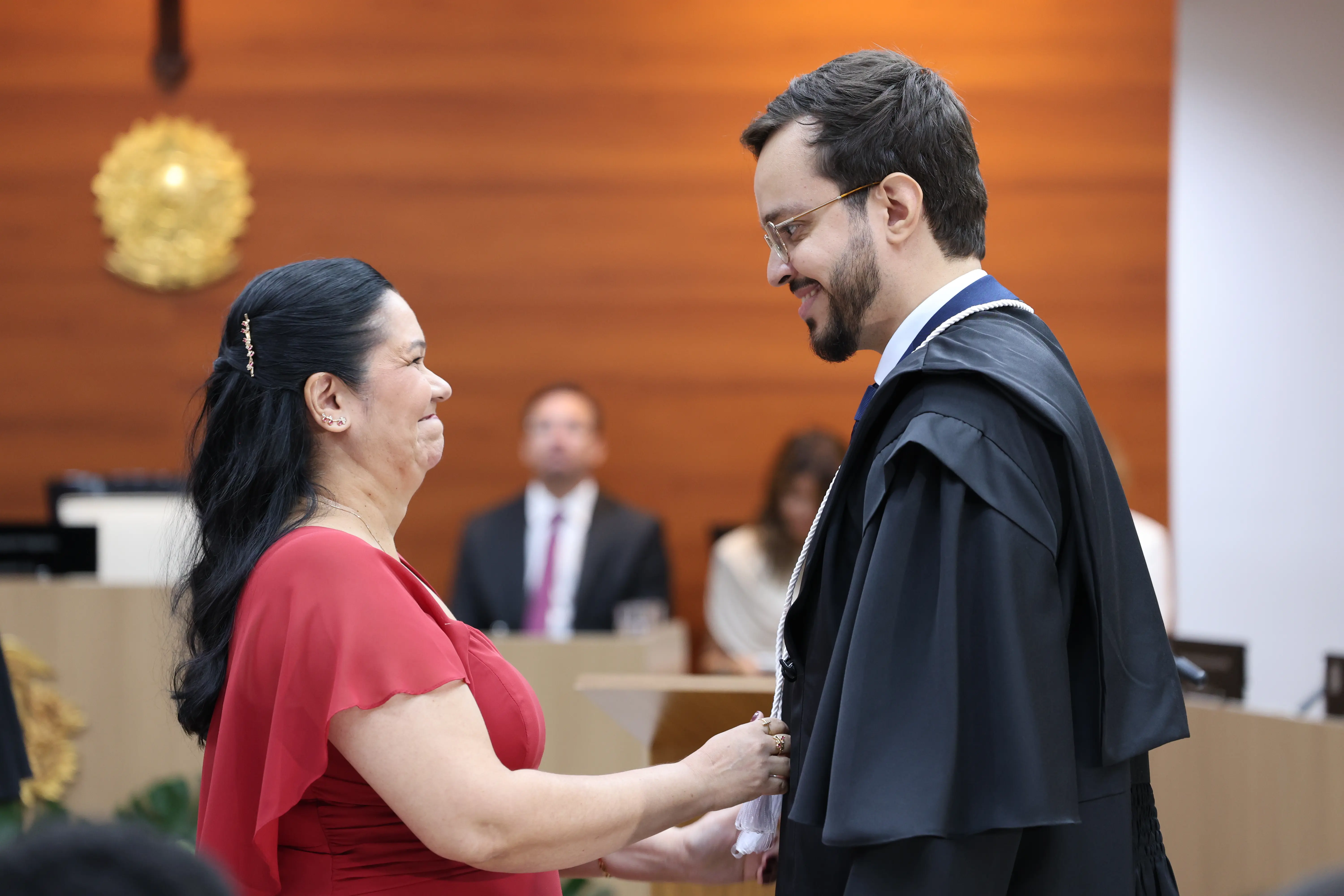 Juiz de toga conversa com familiar durante solenidade, em clima descontraído. O registro foi feito no plenário, com autoridades sentadas e estrutura oficial ao fundo.