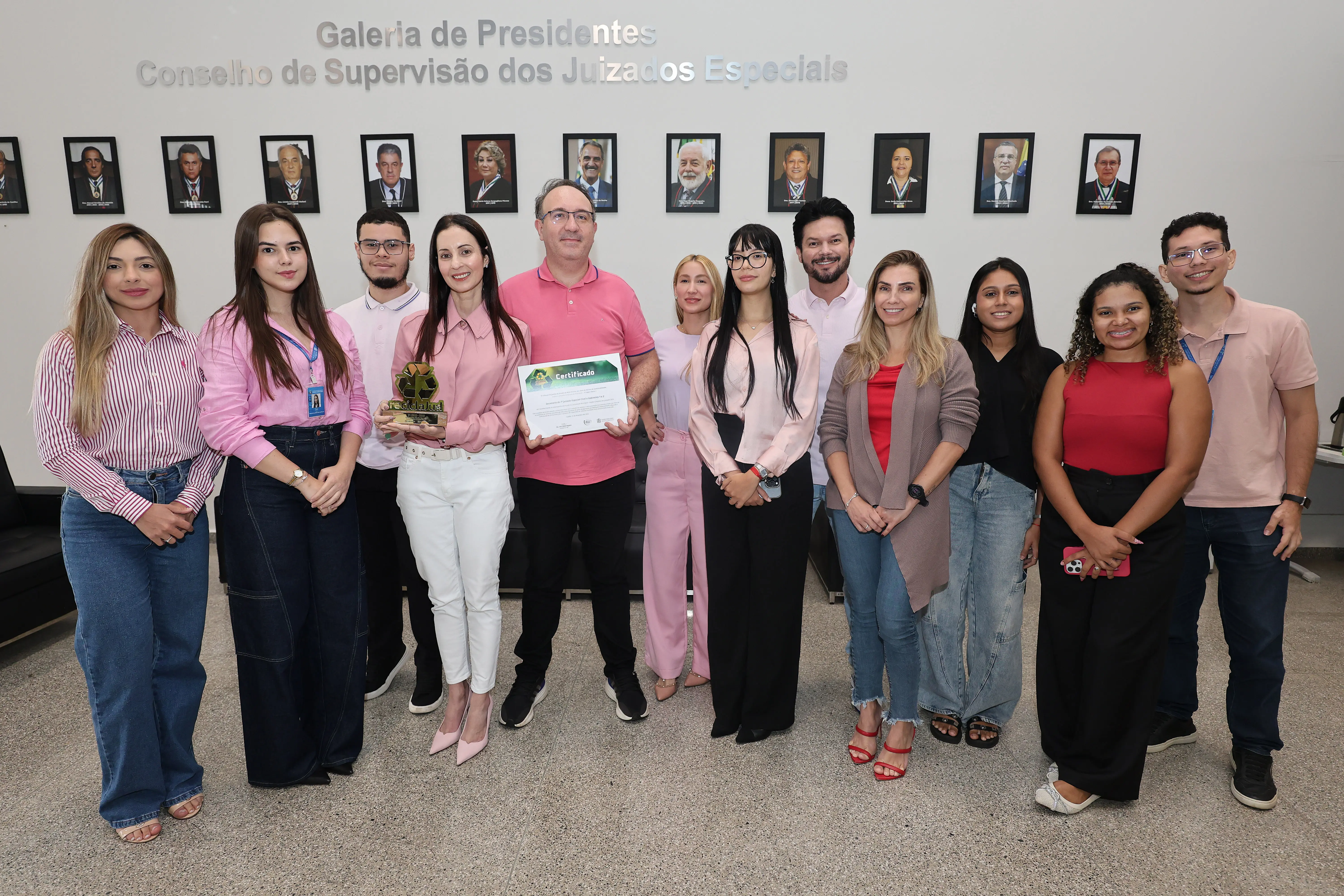 Equipe reunida na Galeria de Presidentes dos Juizados Especiais. O grupo, composto por homens e mulheres jovens e adultos, veste tons de rosa e vermelho. Ao centro, servidores exibem certificado e troféu do ReciclaJud, posando para registro institucional.