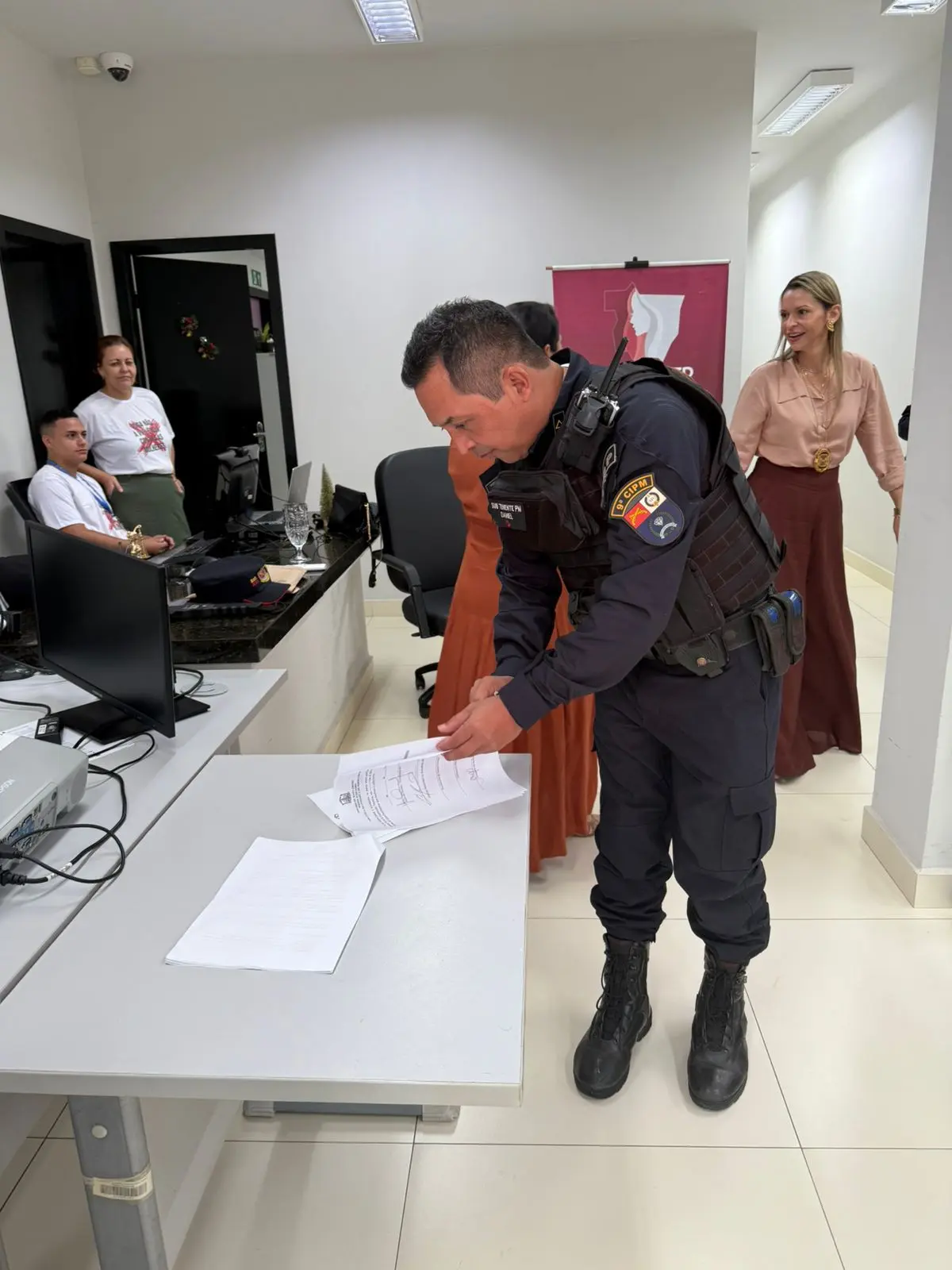 Policial militar fardado assina papéis sobre a mesa, em sala administrativa clara. Ao fundo, servidoras conversam próximas ao banner do CEMULHER. O ambiente mostra a integração dos órgãos no fortalecimento da rede local.