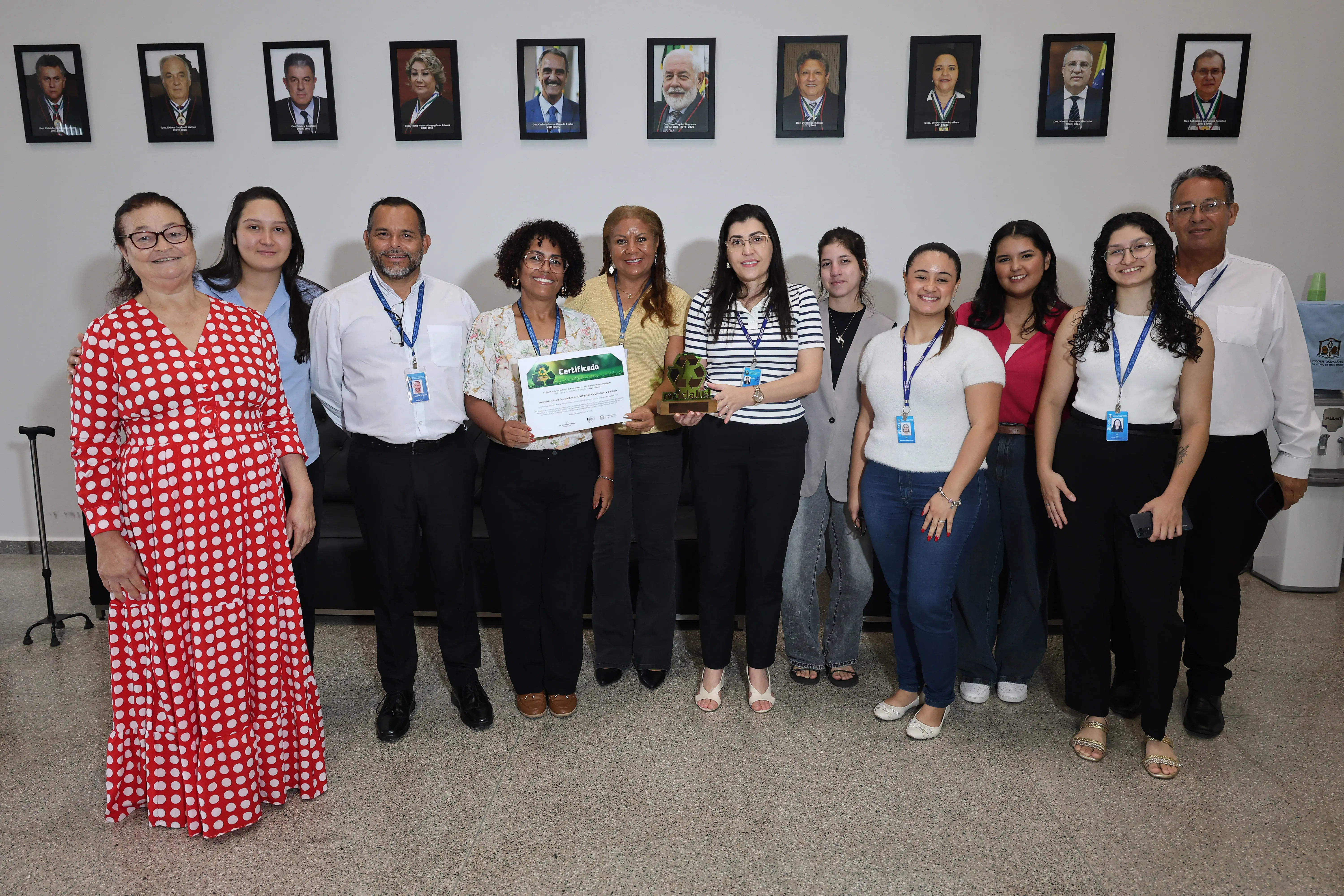 Grupo posicionado em ambiente interno claro, diante da parede com fotos oficiais de magistrados. Servidores do TJMT, homens e mulheres de diferentes idades, posam sorrindo, alguns segurando certificado e troféu do ReciclaJud.