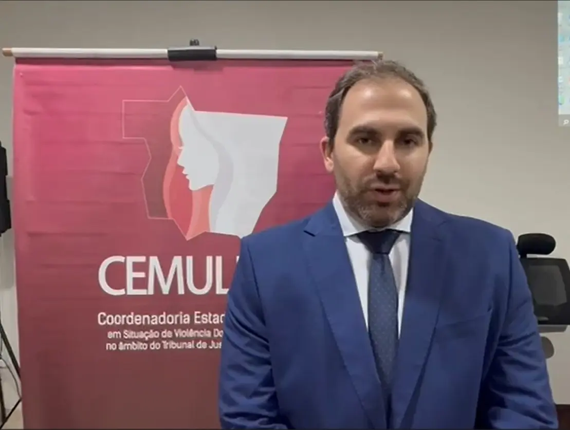 Juiz Daniel Campos é um homem de terno azul e gravata escura aparece diante do banner do CEMULHER. Ele olha direto para a câmera em ambiente interno, com equipamentos e cadeiras ao fundo, durante evento oficial em Nobres.