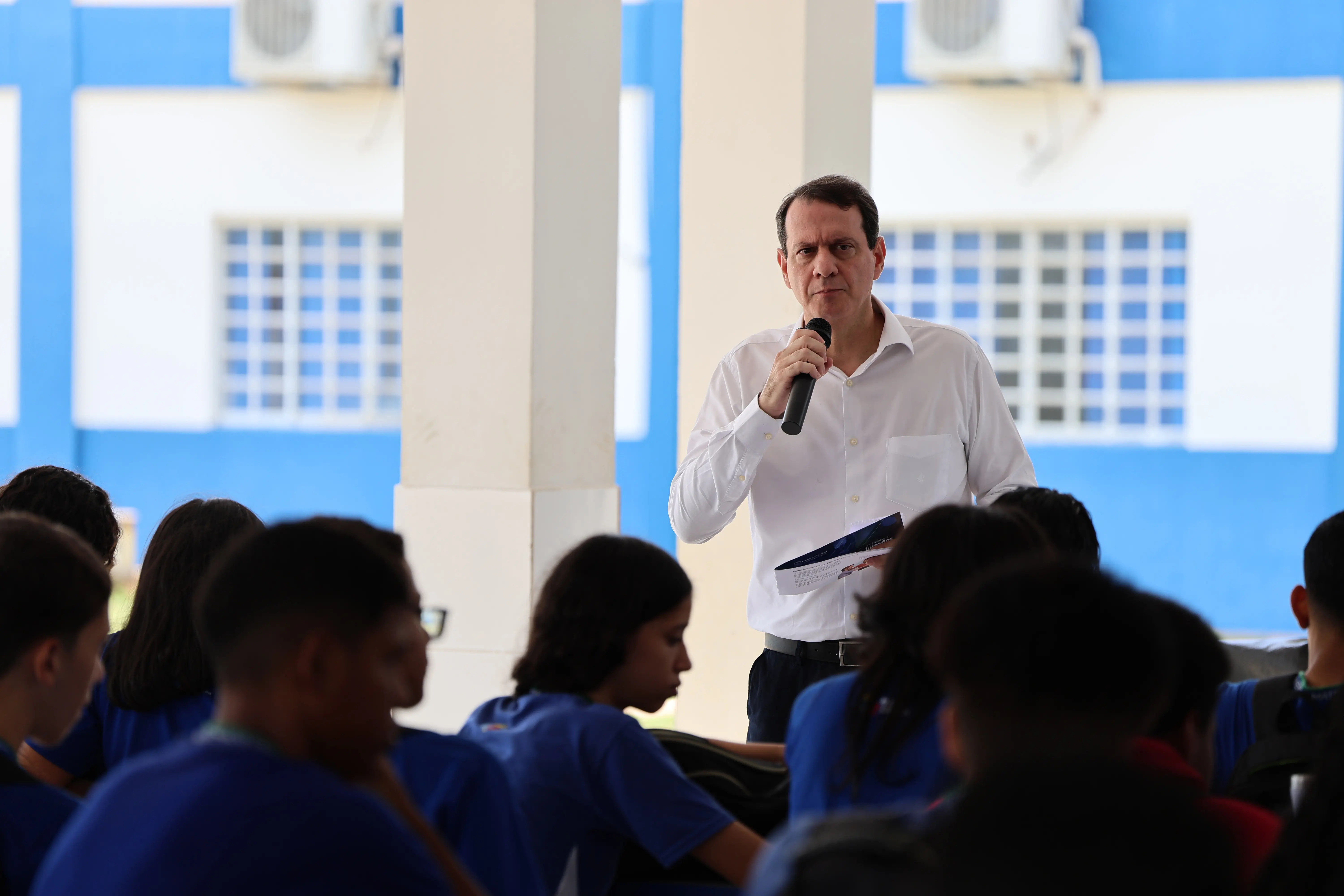 Foto que mostra o palestrante Neifi Feguri, do projeto Nosso Judiciário, falando ao microfone, diante de dezenas de alunos de uma escola estadual.