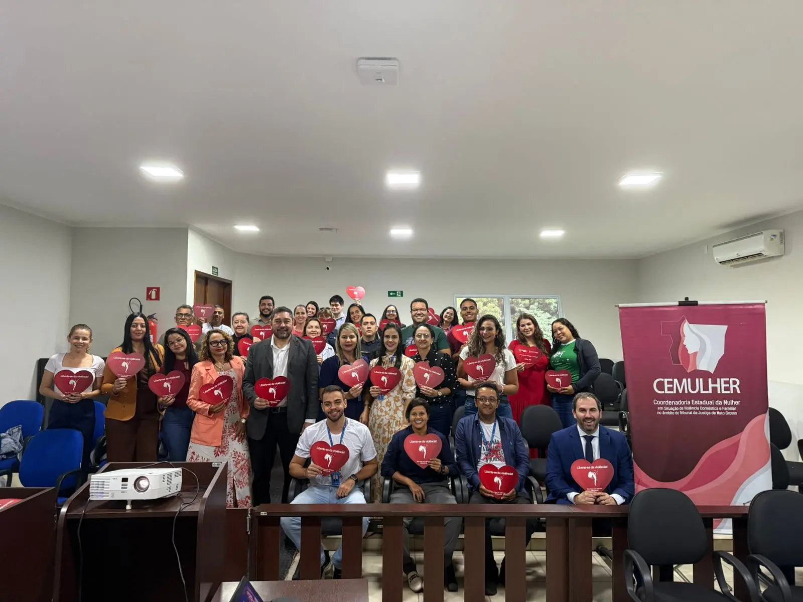 Grupo numeroso posa em auditório do fórum, segurando corações vermelhos do CEMULHER. Homens e mulheres de diferentes órgãos formam a rede, com o banner institucional ao lado e ambiente claro ao fundo.
