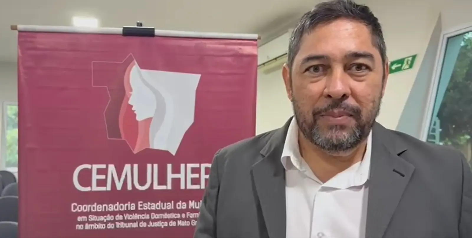 Advogado João Balbino é um homem com barba e cabelo curto, usando terno escuro e camisa branca, aparece diante do banner do CEMULHER. Ele olha para a câmera em ambiente interno claro, com cadeiras e janelas ao fundo.