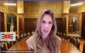 A foto apresenta a juíza Vanessa Carolina Fernandes Ferrari em uma videochamada, com cabelos longos e castanhos. O fundo é notável, mostrando o que parece ser uma sala de reuniões ou um tribunal de época, com uma longa mesa de couro. Há grandes pinturas a óleo, possivelmente de figuras históricas