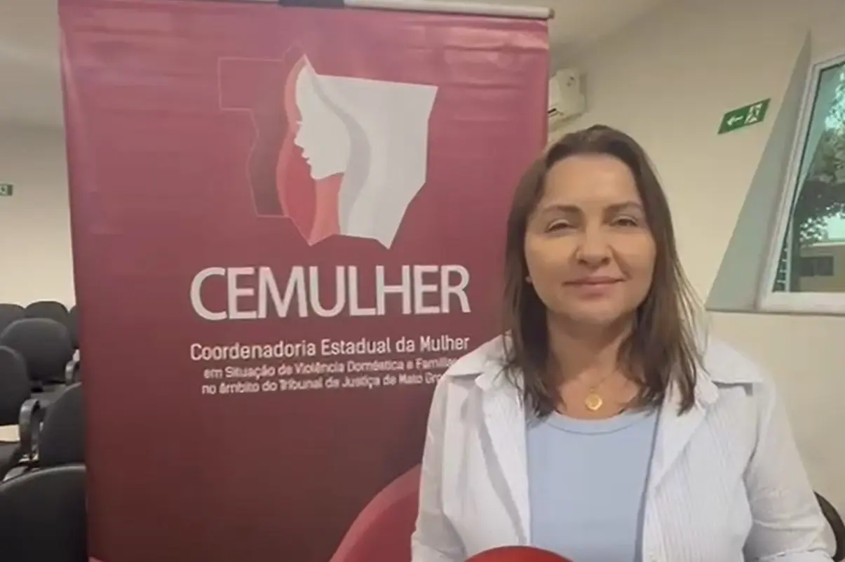 Secretária de Assistência Social Rosemari Camilo é uma mulher de cabelos médios e lisos, veste blusa clara e segura material do evento. Ela posa ao lado do banner do CEMULHER, em sala iluminada com janelas e cadeiras ao fundo.