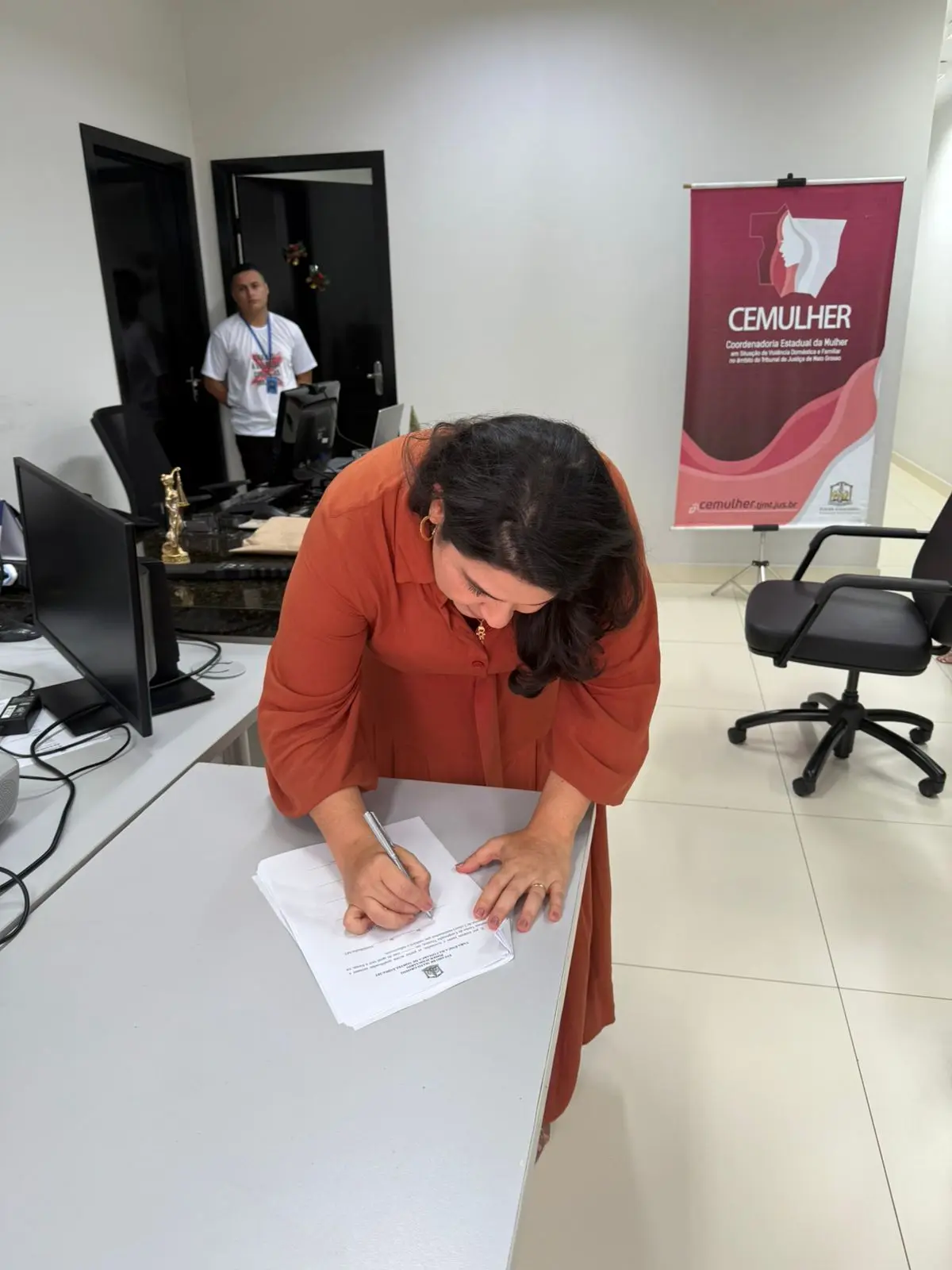 Juíza Lorena Malhado usa vestido terracota assina documentos sobre a mesa em sala clara, com computadores ao fundo. Outra servidora observa ao lado da porta, enquanto banner do CEMULHER indica o caráter institucional do evento.