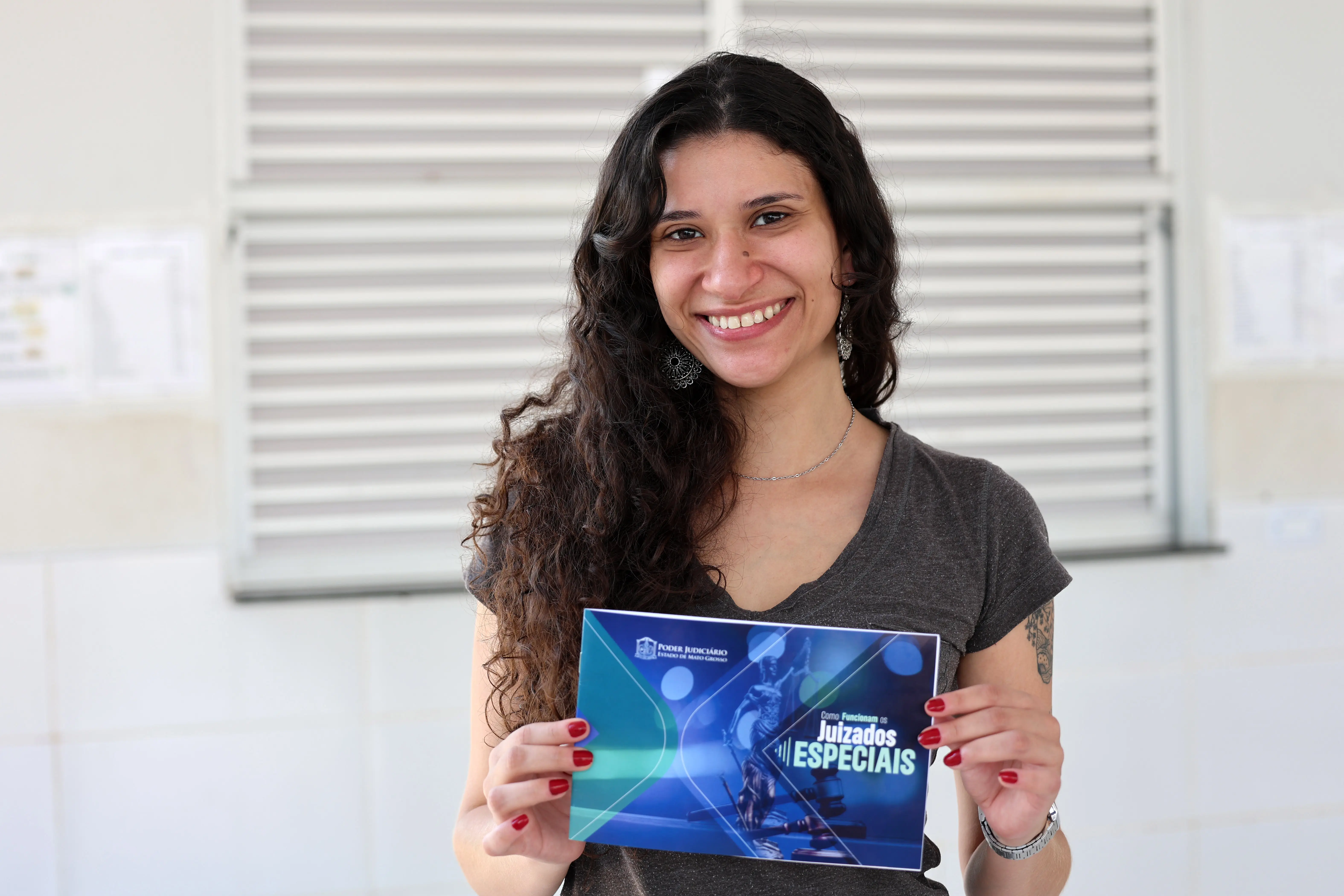Professora Amanda Gouveia sorri para a foto, segurando a cartilha do projeto Nosso Judiciário. Ela é uma mulher jovem, branca, magra, com longos cabelos cacheados e castanhos, usando camiseta cinza. 
