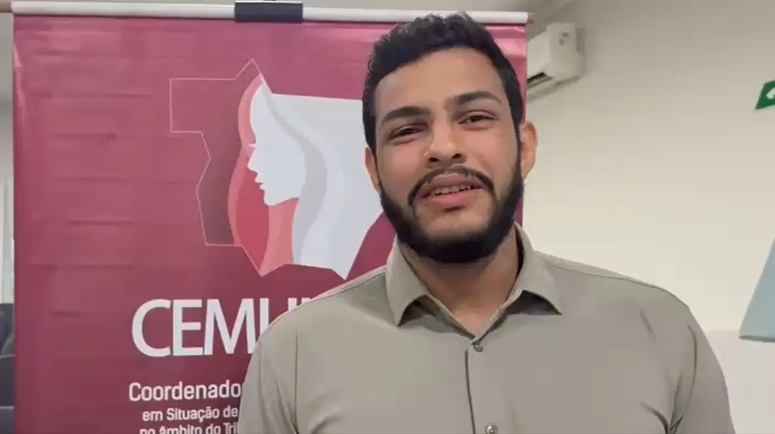 Kadrialson Ribeiro é um homem jovem com barba aparada e camisa clara sorri levemente em frente ao banner do CEMULHER. A sala é iluminada, com paredes brancas e cadeiras ao fundo, reforçando o clima institucional do evento