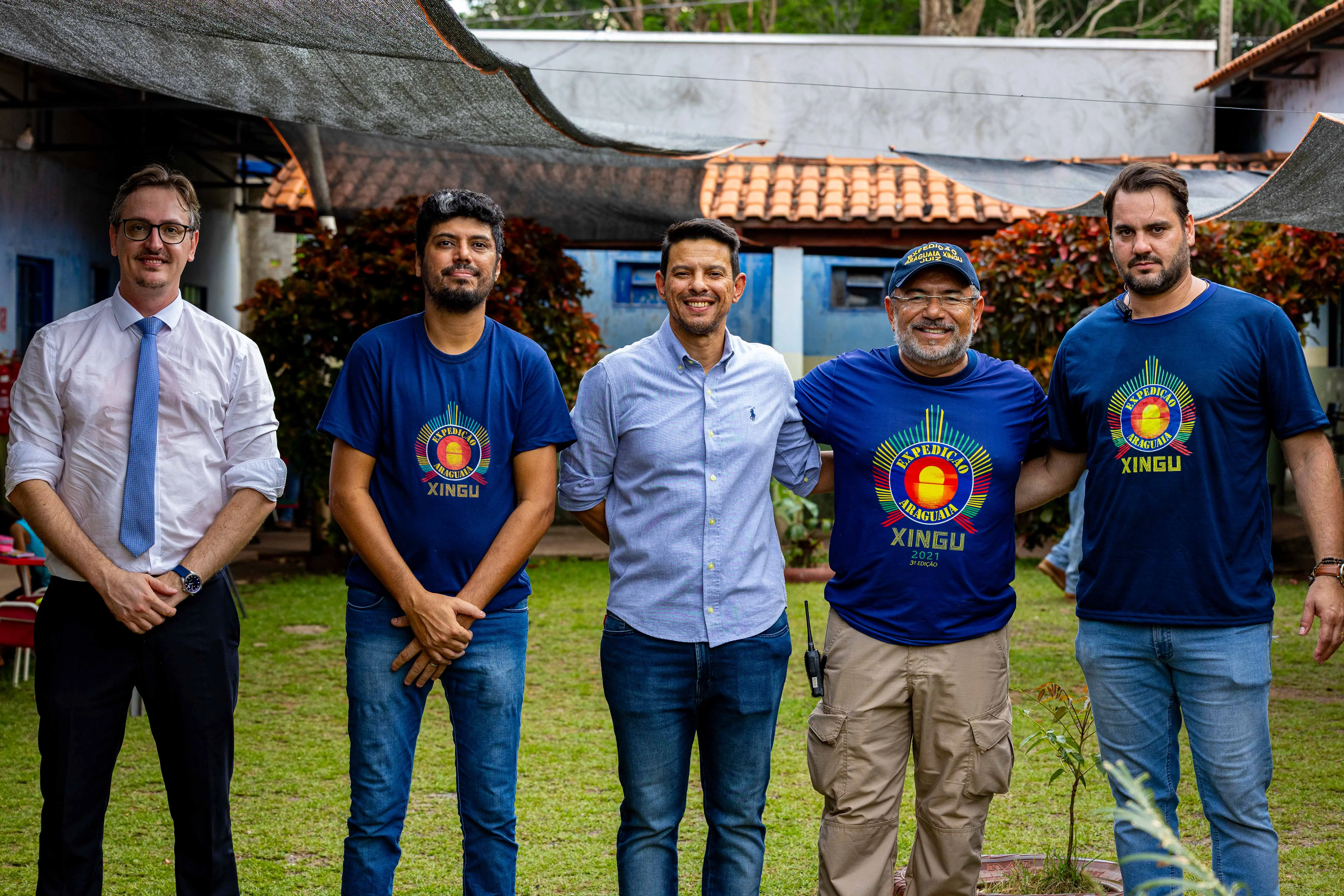 Cinco homens de pé, sorrindo, em uma foto ao ar livre. Um usa gravata e os demais, roupas casuais, incluindo camisetas azuis de evento e boné.