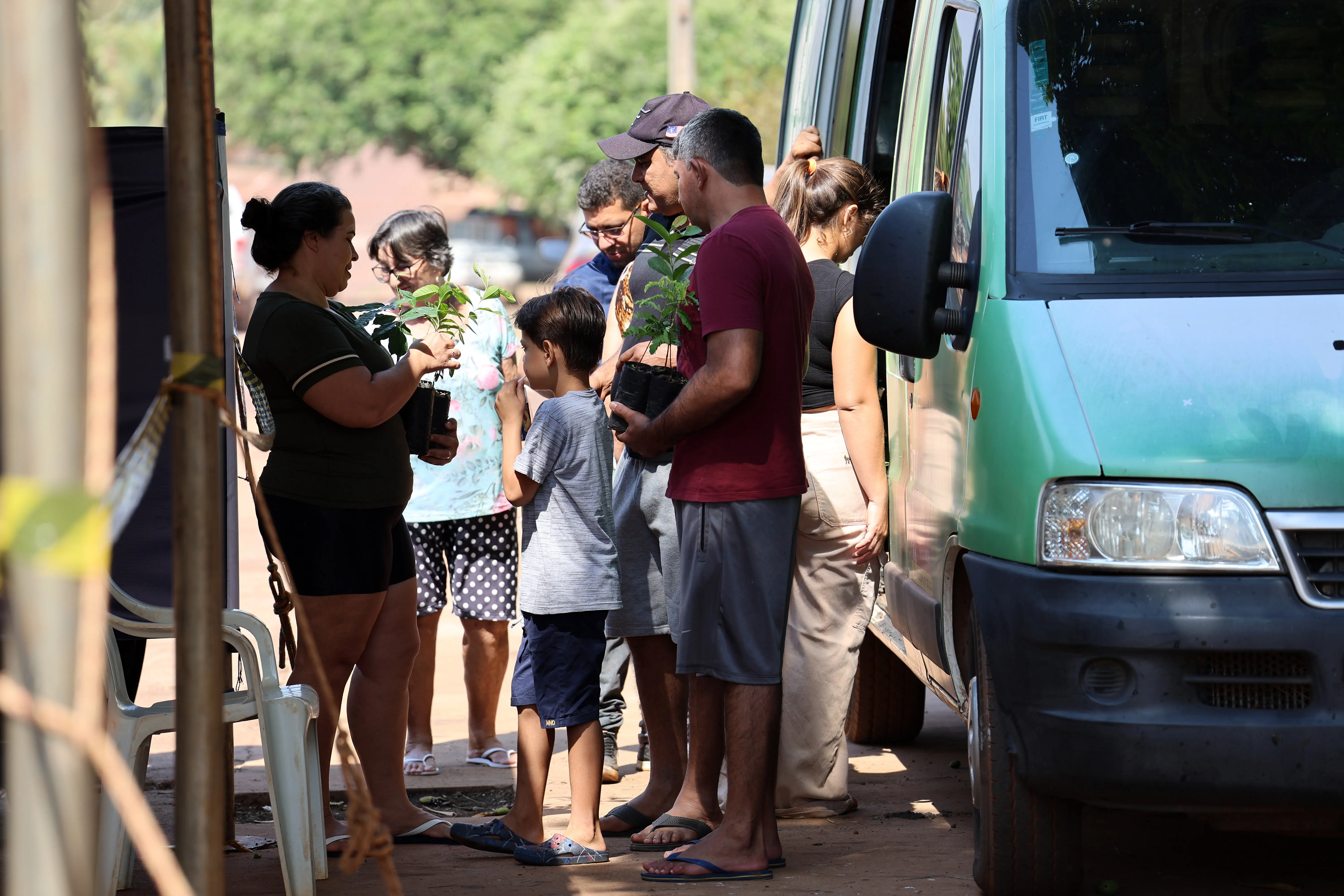 Famílias formam fila para receber mudas ao lado da van do Verde Novo. Adultos e crianças observam curiosos as pequenas plantas, demonstrando interesse e envolvimento ambiental.