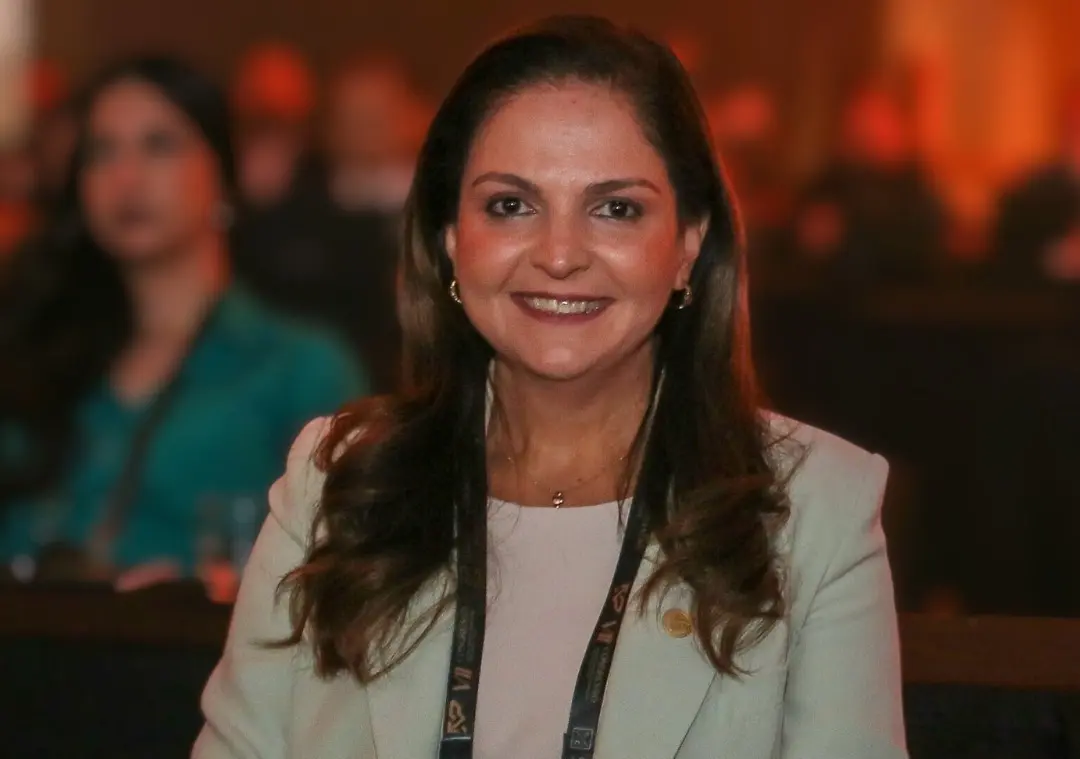 Presidente da OAB-MT, Gisela Cardoso está sentada em evento formal, usando blazer claro e crachá. Ao fundo, outras pessoas também estão sentadas. O ambiente parece ser uma sala de conferência ou auditório, com atmosfera institucional.