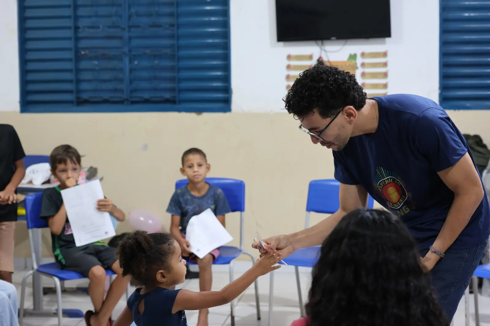 Gestor do Cejusc entrega doce a uma menina pequena durante o Círculo de Paz. Crianças observam e seguram desenhos e balões, em clima de carinho e atenção.