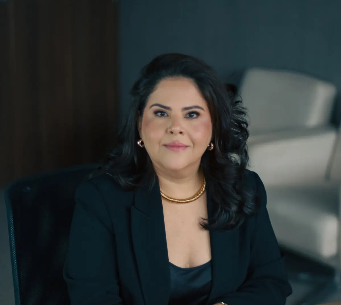 Pessoa em escritório moderno, com blazer escuro e acessórios dourados. O ambiente tem cadeiras claras e paredes escuras, transmitindo sofisticação e profissionalismo. A composição sugere contexto jurídico ou institucional.