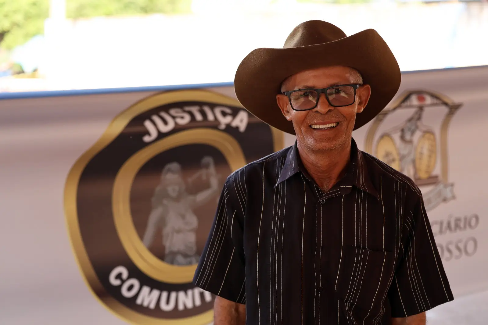 Homem de chapéu sorri diante do banner da Justiça Comunitária, com logotipo ao fundo. Ele representa o público atendido pela Expedição Araguaia-Xingu