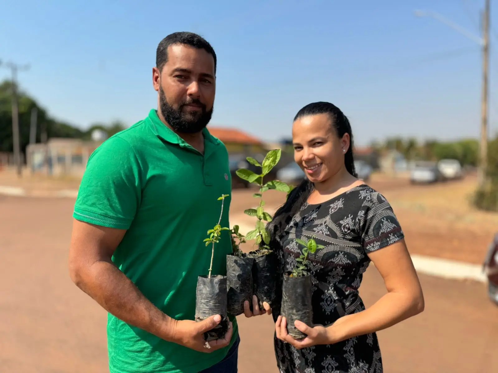 Casal sorri ao segurar pequenas mudas em uma rua ensolarada. Ele veste camisa verde e ela, vestido estampado preto. A cena transmite alegria e envolvimento com ações ambientais e comunitárias.