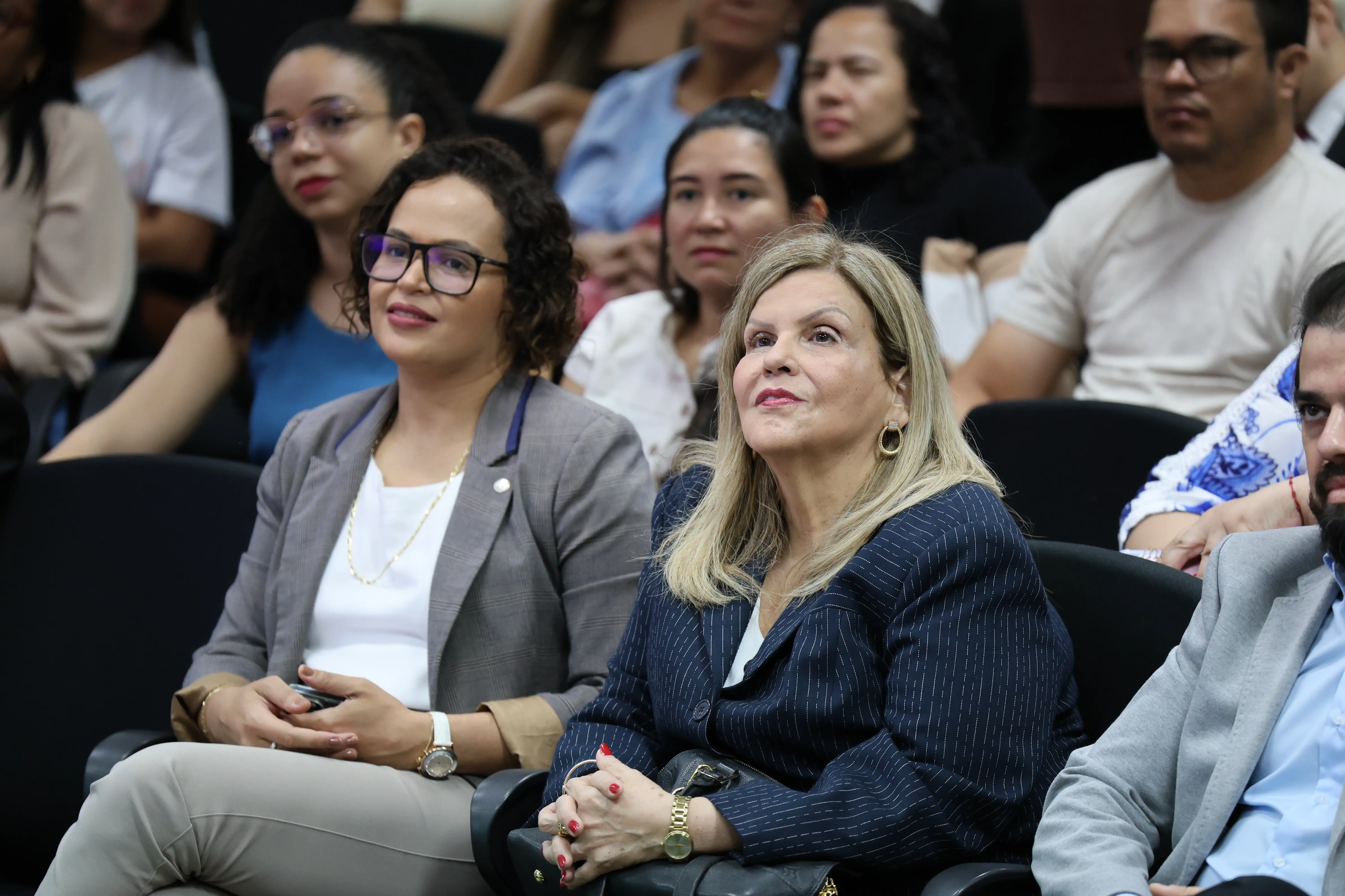A imagem mostra desembargadora Nilza e um grupo de pessoas sentadas em ambiente de conferência ou seminário. Estão vestidas de forma casual ou profissional e parecem atentas ao que ocorre fora do enquadramento, indicando participação ativa no evento.