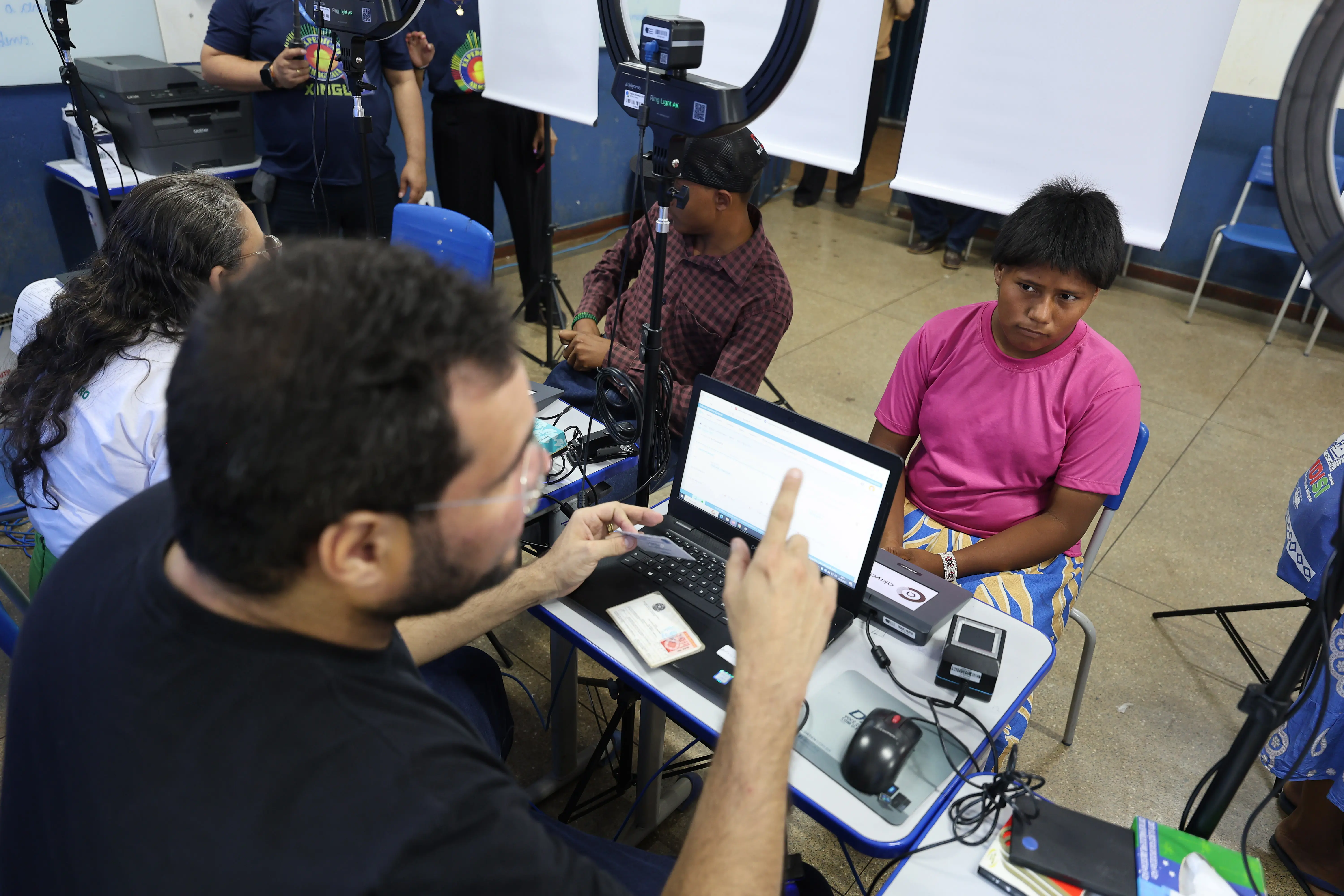 Em sala de atendimento, profissional do Tribunal Regional Eleitoral conversa com jovem indígena Xavante vestida com camiseta rosa, enquanto realiza cadastro em computador. O ambiente tem mesas, cadeiras e equipamentos de registro.