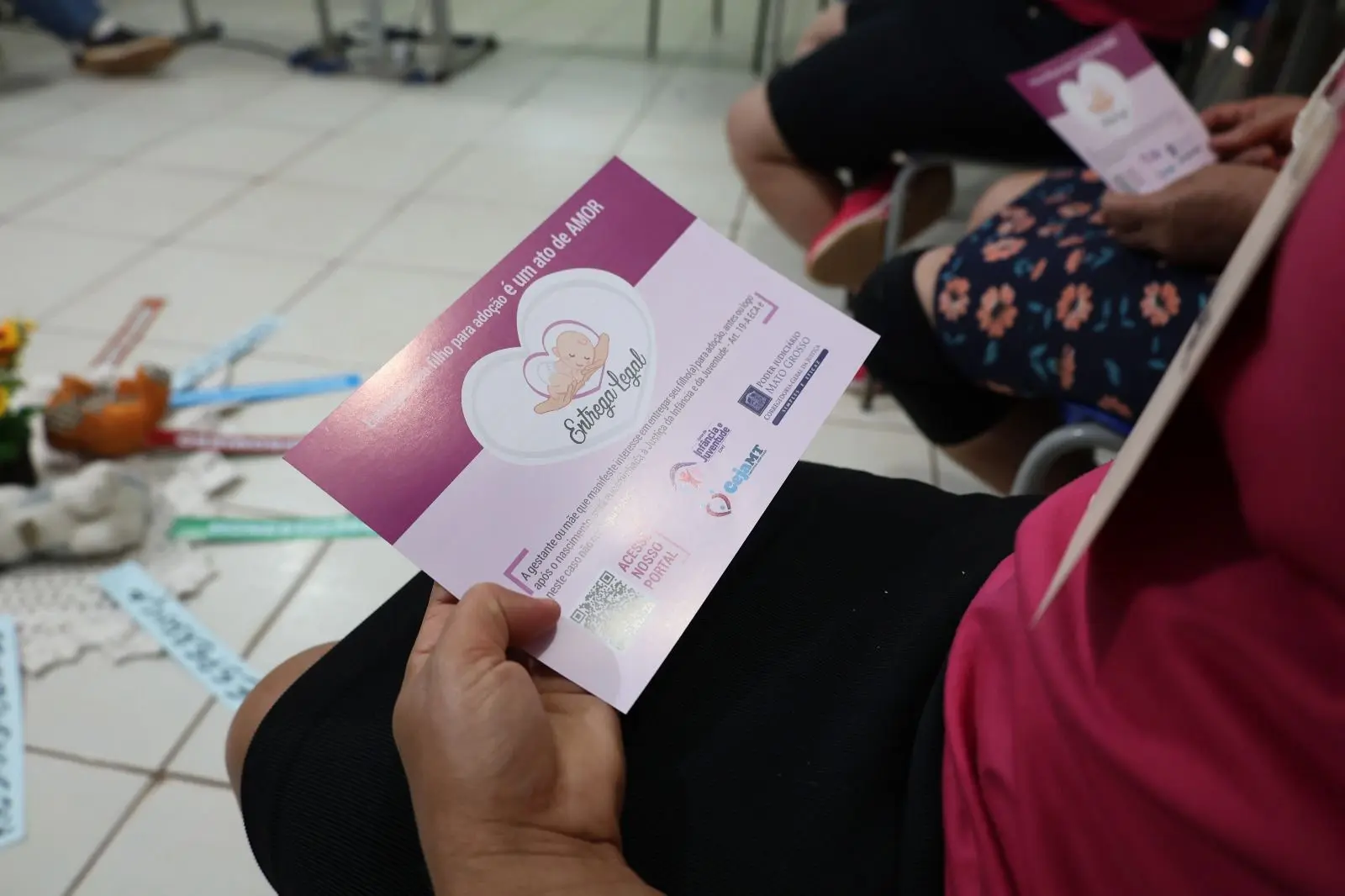 Pessoa segura um folder cor-de-rosa com a frase “Entregar um filho para adoção é um ato de amor”. No fundo, há flores e faixas com palavras como “honestidade” e “esperança”, compondo um cenário acolhedor de reflexão.