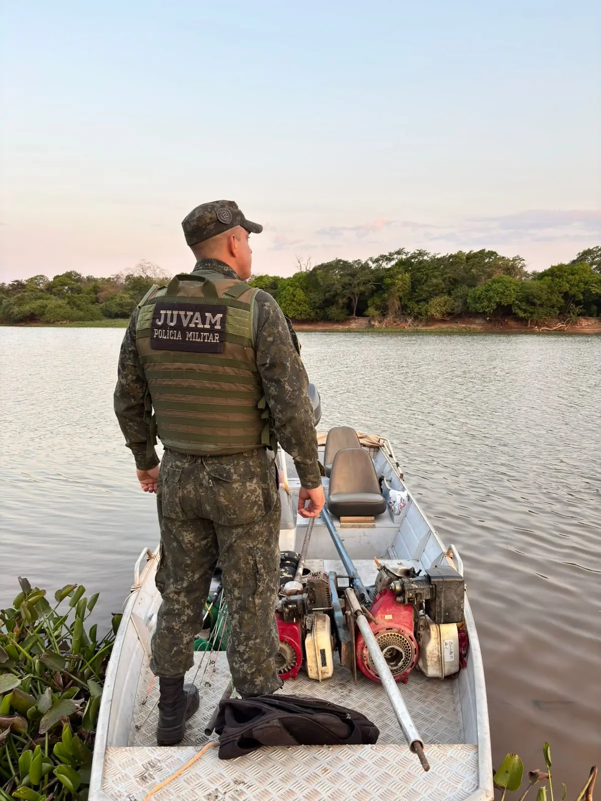 Agente do Juvam, da Polcia Militar Ambiental, observa o rio em misso de fiscalizao. Ele est em um barco metlico com motores apreendidos e vegetao aqutica ao redor, sob luz suave do entardecer.