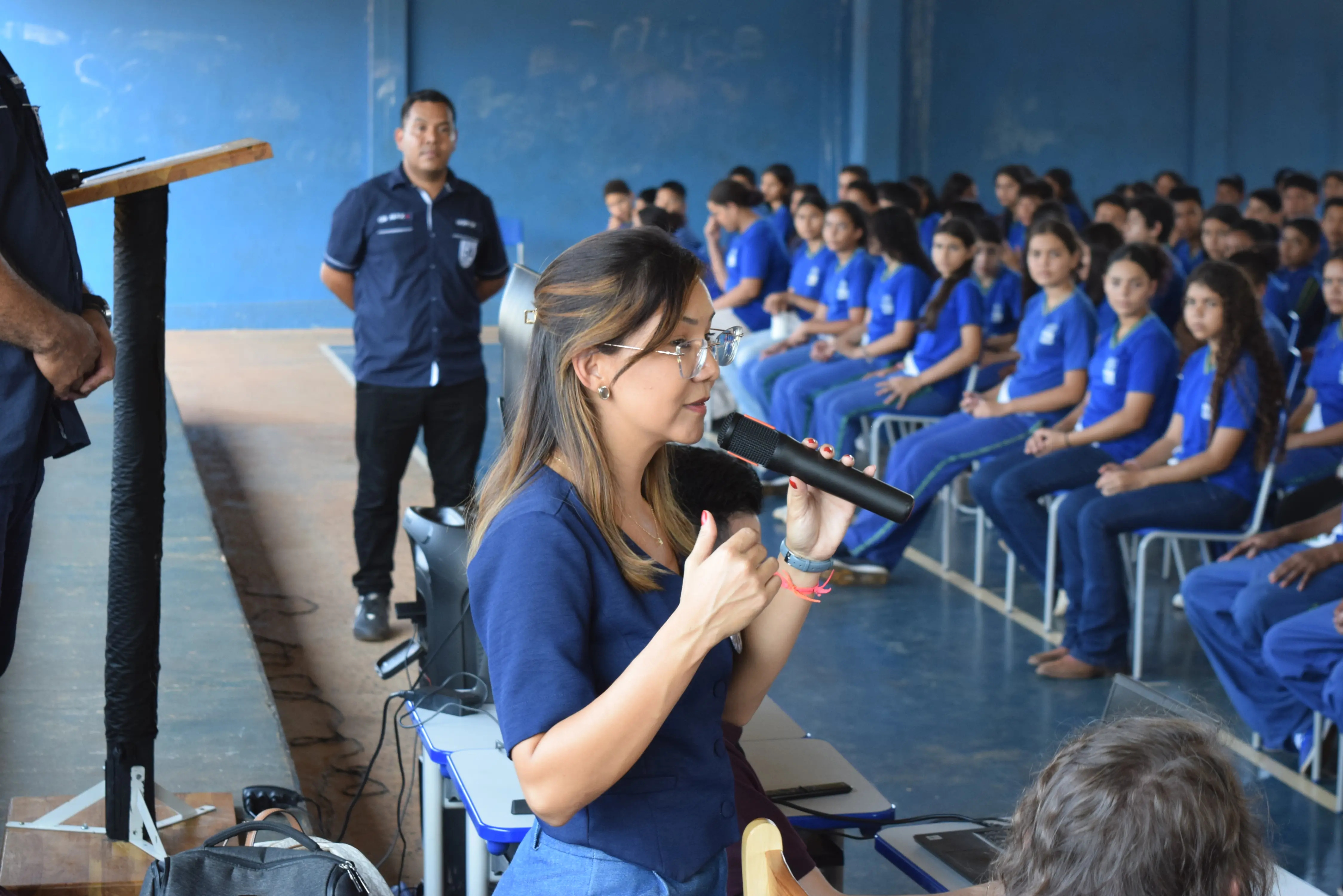 Mulher de óculos e blusa azul fala ao microfone em plano médio. À direita, diversos estudantes de uniforme azul assistem sentados na arquibancada de uma quadra coberta.