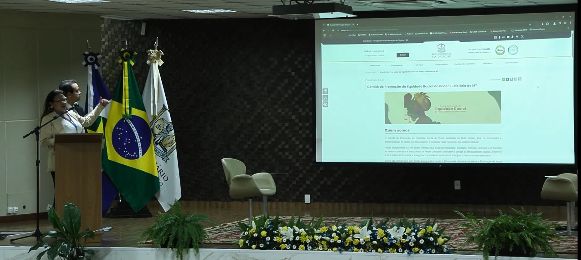 Foto em plano aberto que mostra o juiz Emerson Cajango e a servidora Kelly Assumpção no palco do auditório, apresentando a página do Comitê de Equidade Racial, que aparece no telão. 
