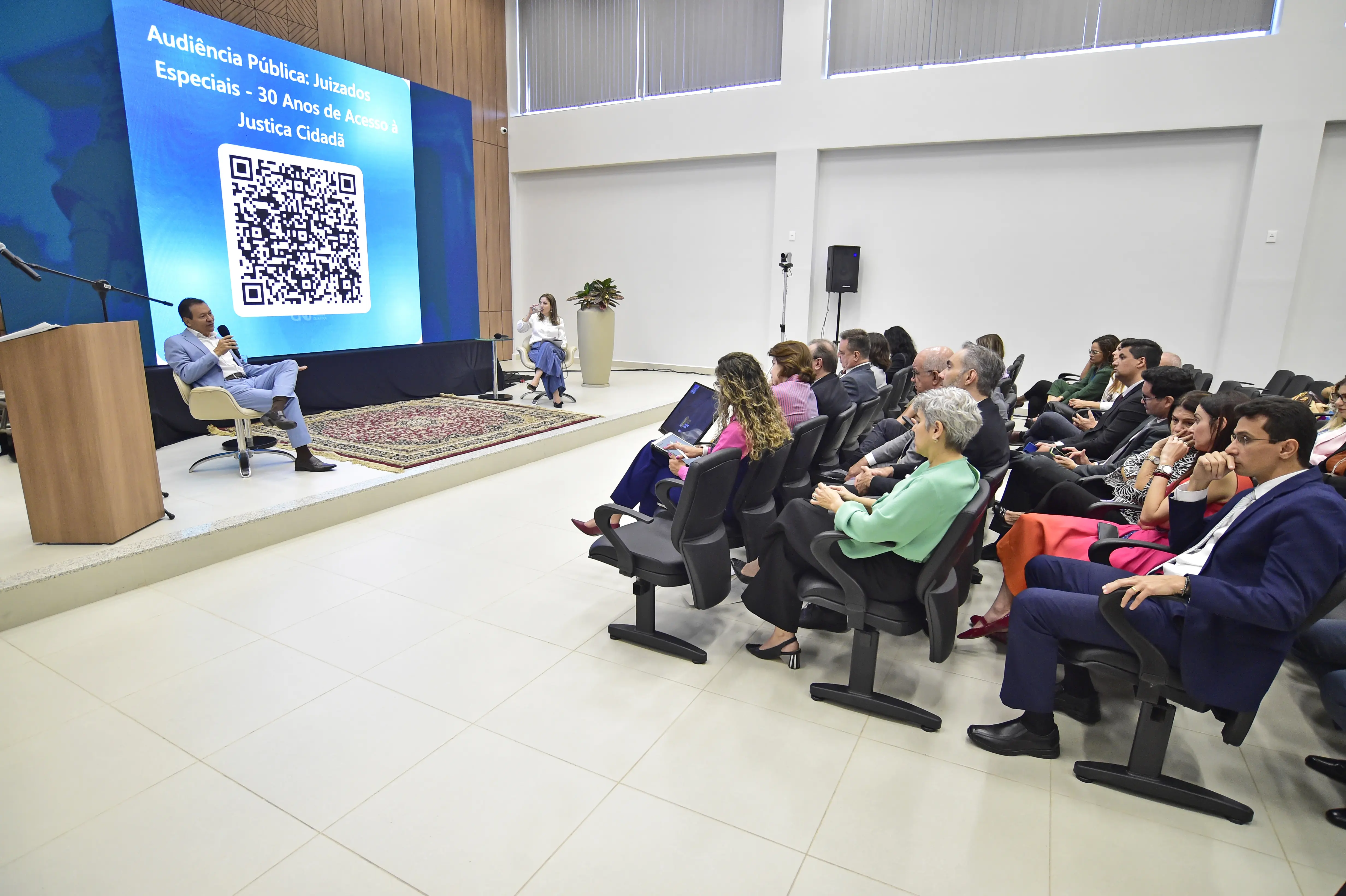 Auditório com um palestrante sentado em poltrona, falando ao microfone. Um grande telão exibe um QR Code e o tema 'Audiência Pública: Juizados Especiais'. A plateia está sentada em filas, atenta