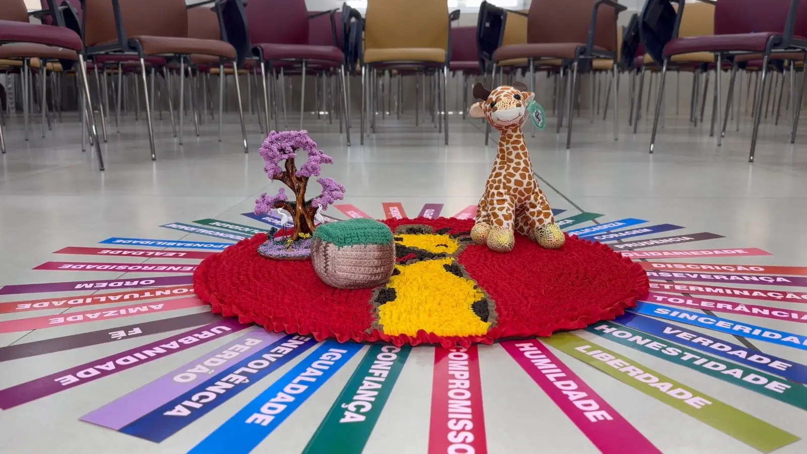 No centro de um salão, um círculo colorido feito com tecidos e flores representa o Círculo de Paz. No meio, estão figuras de animais e uma vela acesa, simbolizando harmonia e união entre as pessoas.