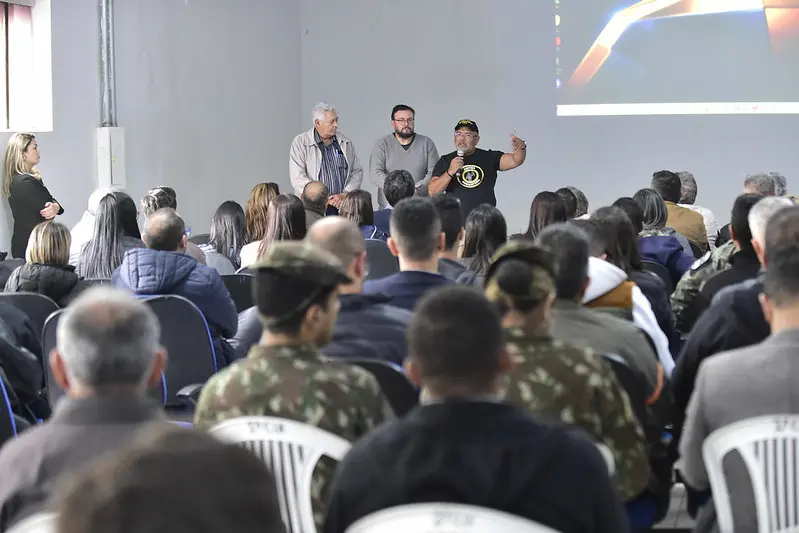 Em um auditório do Batalhão do Exército, dezenas de pessoas estão sentadas, de costas na foto. À frente, o juiz José Antônio Bezerra Filho fala ao microfone, ao lado do juiz de Cáceres e do secretário de Assuntos Estratégicos de Cáceres.