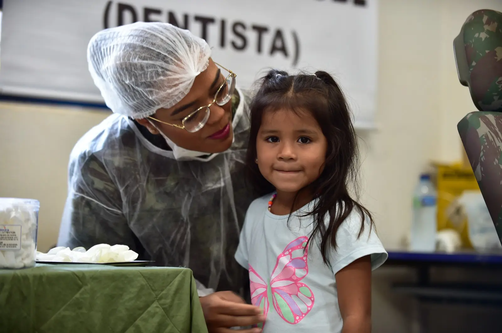 Dentista militar atende menina pequena em consultório. A criança sorri timidamente para a câmera, enquanto a dentista, com touca e jaleco, a observa com atenção e cuidado.
