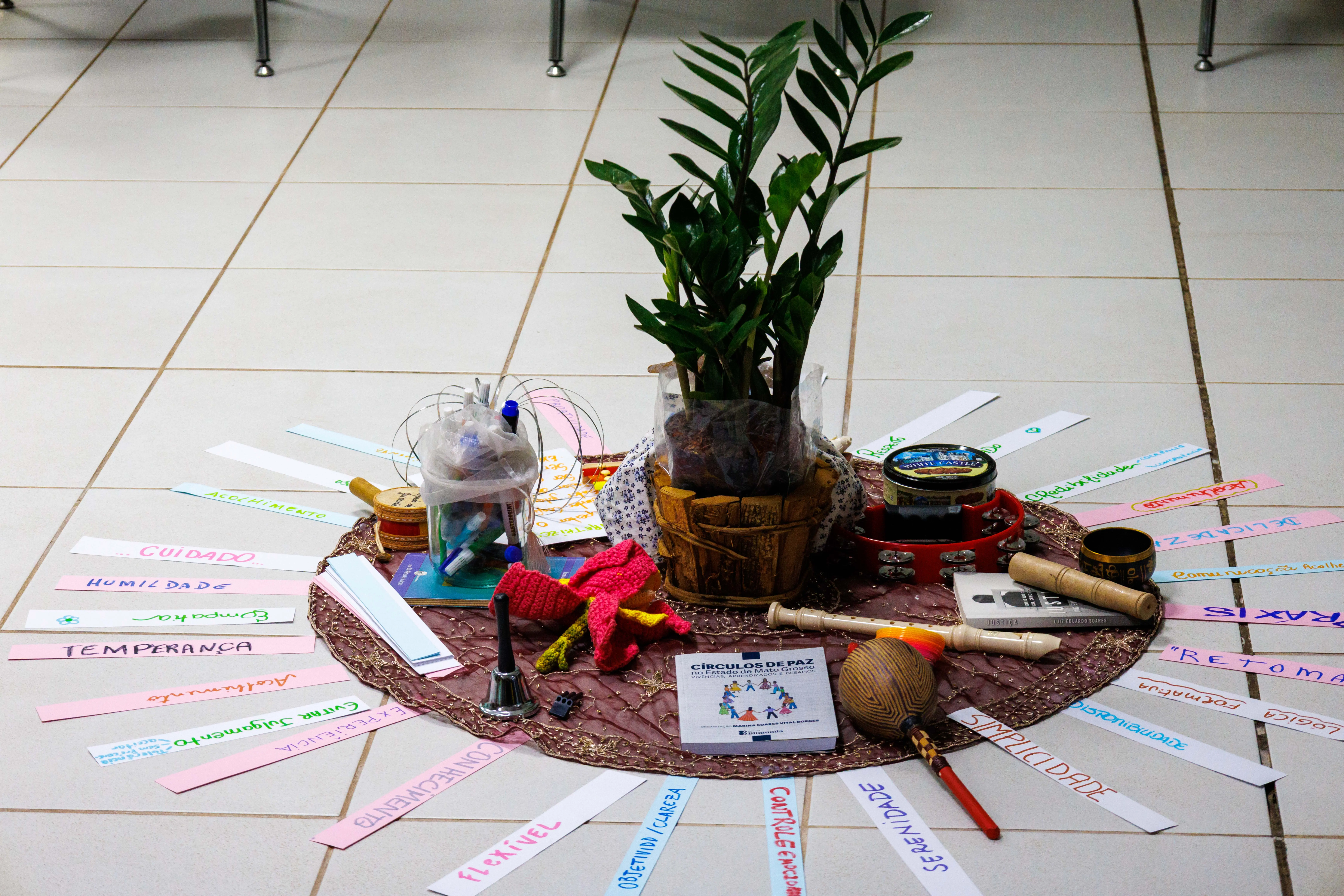 Círculo de objetos coloridos no chão exibe planta em vaso, instrumentos musicais, cartazes com palavras como “cuidado” e “humildade” e cartilha de círculos de paz, simbolizando valores de convivência.