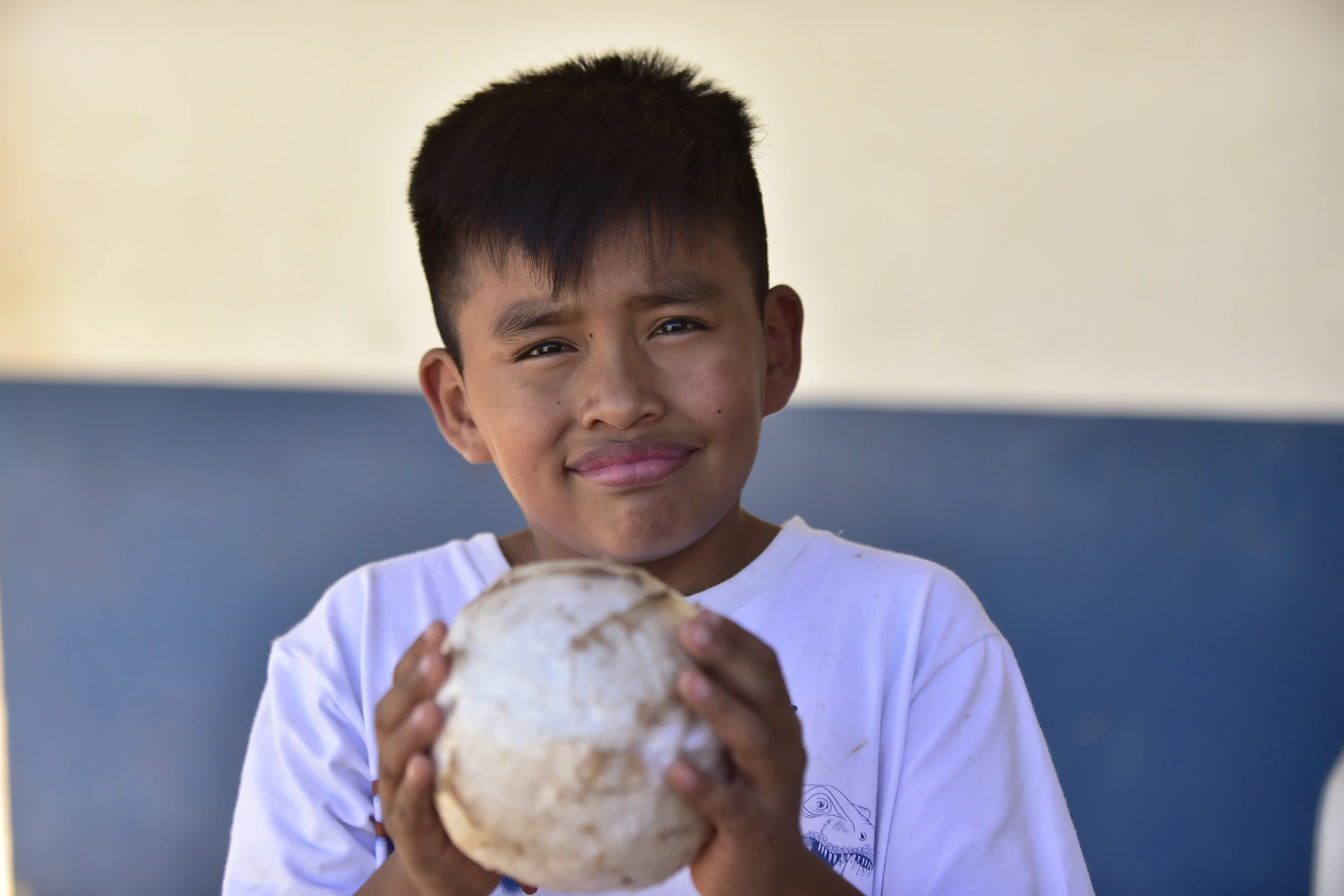 Menino negro, de cabelos lisos e pretos, olhos escuros e vestindo camiseta branca sorri para a foto. Nas mãos, ele segura uma bola de futebol.
