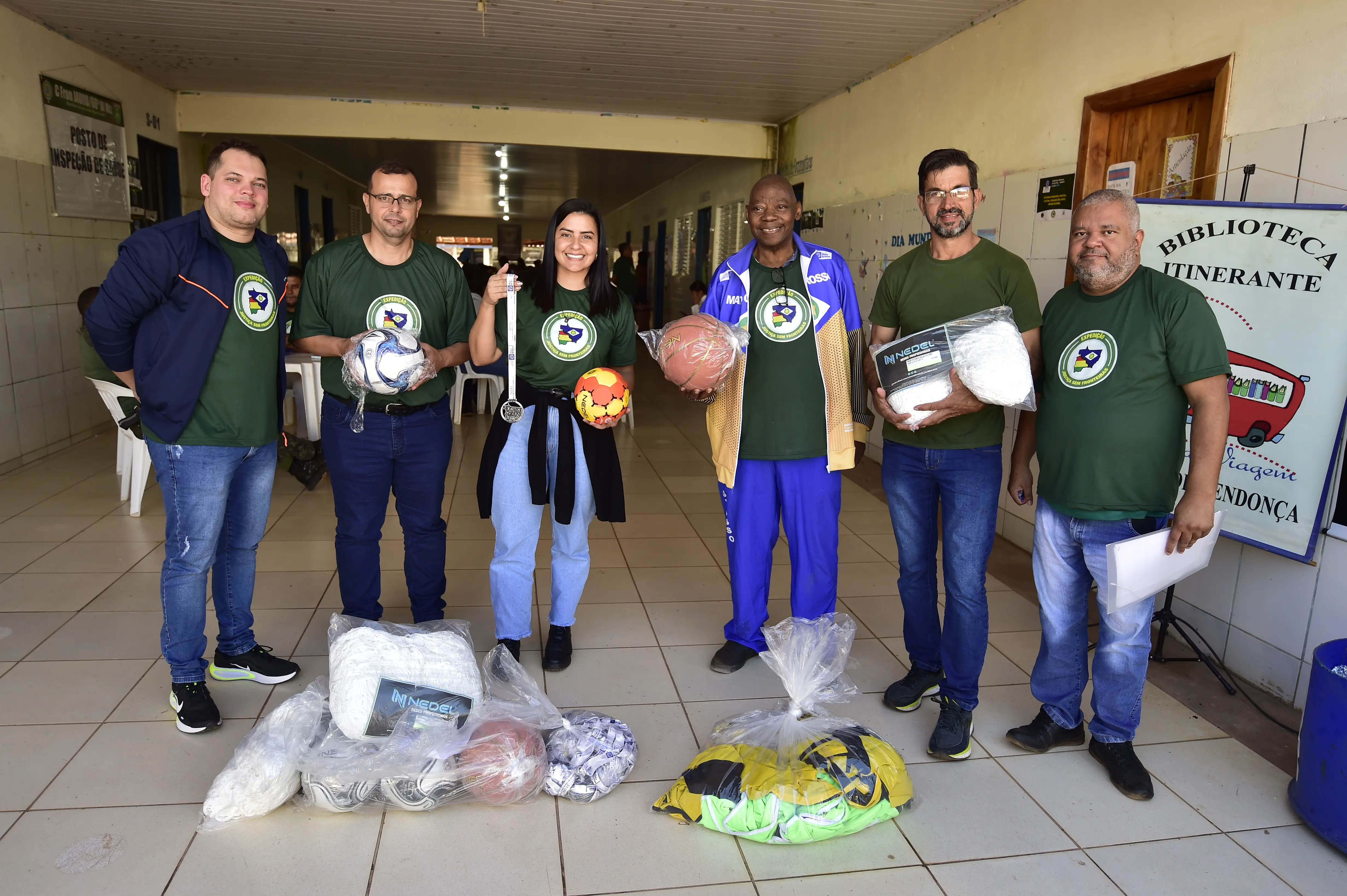 Servidores da Secretaria de Estado de Esporte e prefeito de Porto Esperidião posam sorrindo, durante entrega de kits esportivos. Eles estão no pátio de uma escola e usam a camiseta verde da Justiça Sem Fronteiras.